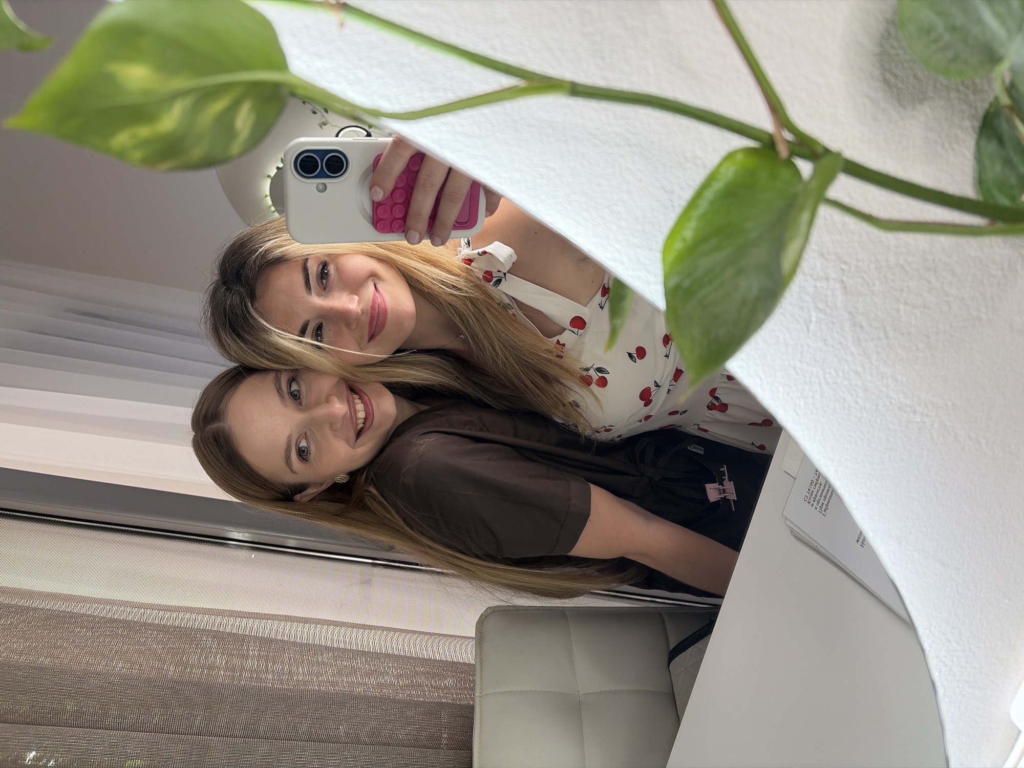 This image is a mirror selfie taken indoors in Budva, Montenegro, featuring two young adult women. The foreground on the right is partially obscured by an out-of-focus green leafy plant with visible stems.
The woman on the left possesses long, straight brown hair with lighter highlights. She wears a dark brown, short-sleeved top or robe, cinched at the waist, and gold hoop earrings. She is smiling broadly and looking towards the right, with her left arm positioned around the second woman.
The woman on the right has long, wavy blonde hair. She is wearing a white top or dress adorned with a repeating red cherry pattern and features tie straps. A small, simple necklace is visible around her neck. She holds a white smartphone, equipped with a blue dual-camera module and a pink circular pop socket or grip, in her right hand, angled to capture the reflection. She is also smiling, looking directly into the mirror/camera.
The background, visible in the mirror's reflection, includes a white upholstered chair with a tufted back on the far left. Behind the women, windows are covered by a textured brown roller blind or curtain on the left and white vertical blinds or sheer curtains on the right. A white desk or table surface is present in the lower center and left portion of the reflection, where a document with partially legible Cyrillic text is visible. The interior space is well-lit.