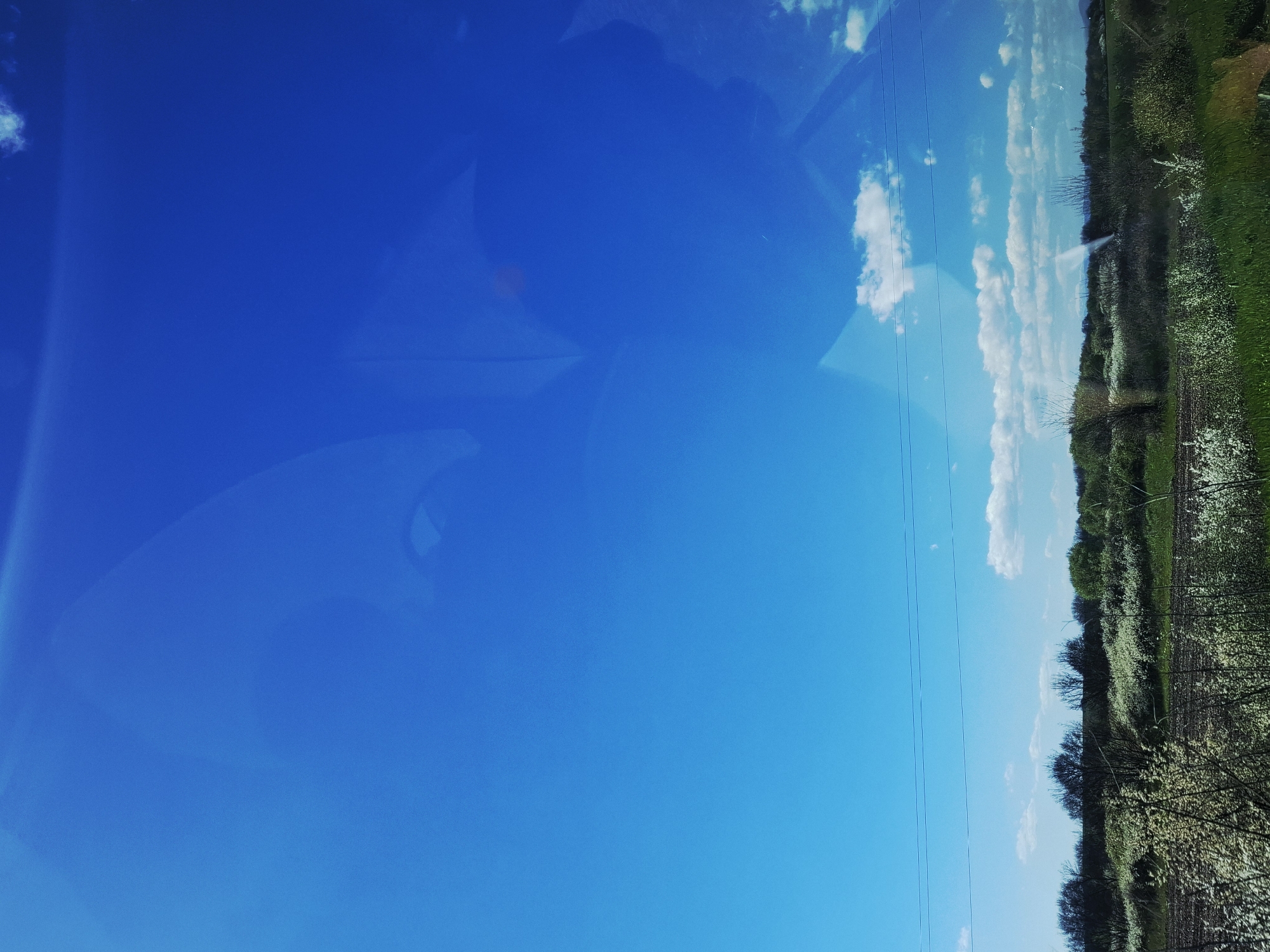The image shows a view of a clear blue sky with some scattered white clouds. In the lower right portion of the image, there is a glimpse of the landscape, including green vegetation, trees with white blossoms, and what appears to be plowed land. The presence of wires suggests the scene may be near some infrastructure. The sky seems to be reflected on a window.