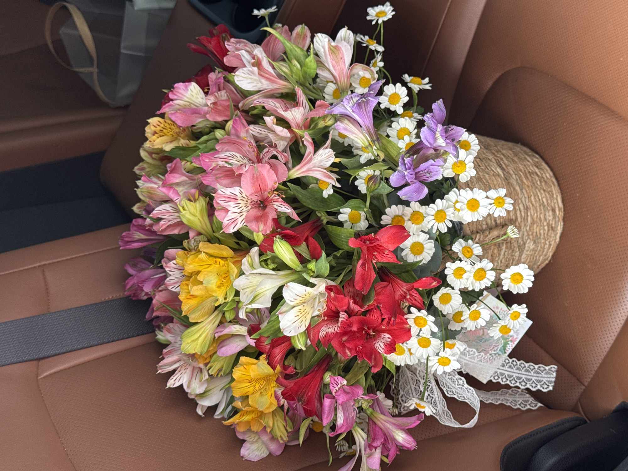 A full shot shows a bouquet of alstroemeria and daisies in a woven basket. The basket is placed on a brown leather car seat, likely in a vehicle in Almaty, Kazakhstan. The bouquet features a variety of colors, including pink, yellow, white, and red alstroemeria, interspersed with white daisies. A white lace ribbon is tied around the basket, with a small card attached. The background shows the interior of a car, with a seatbelt visible. The lighting is natural, suggesting it was taken during the daytime.