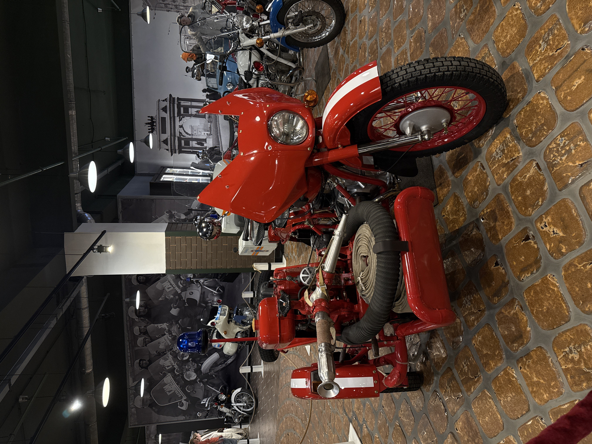 The image depicts the interior of a museum, likely the Arkhangelskoye Estate Museum in Arkhangelskoye, Russia, featuring a collection of vintage motorcycles and sidecars. In the foreground, a striking red motorcycle with a sidecar equipped for firefighting is prominently displayed. This specialized vehicle has a blue flashing light on top, a fire hose coiled neatly, and a nozzle at the front. Behind it, other vintage motorcycles, including a white one with blue accents and a white and black police motorcycle, are visible. The museum's walls are adorned with large black and white murals of historical scenes, including what appears to be a parade of police officers on motorcycles and a view of the Brandenburg Gate. The floor is paved with an intricate pattern of cobblestones. The lighting suggests an indoor exhibition space with overhead track lighting and ambient illumination. No people are actively engaged with the exhibits in the immediate foreground.