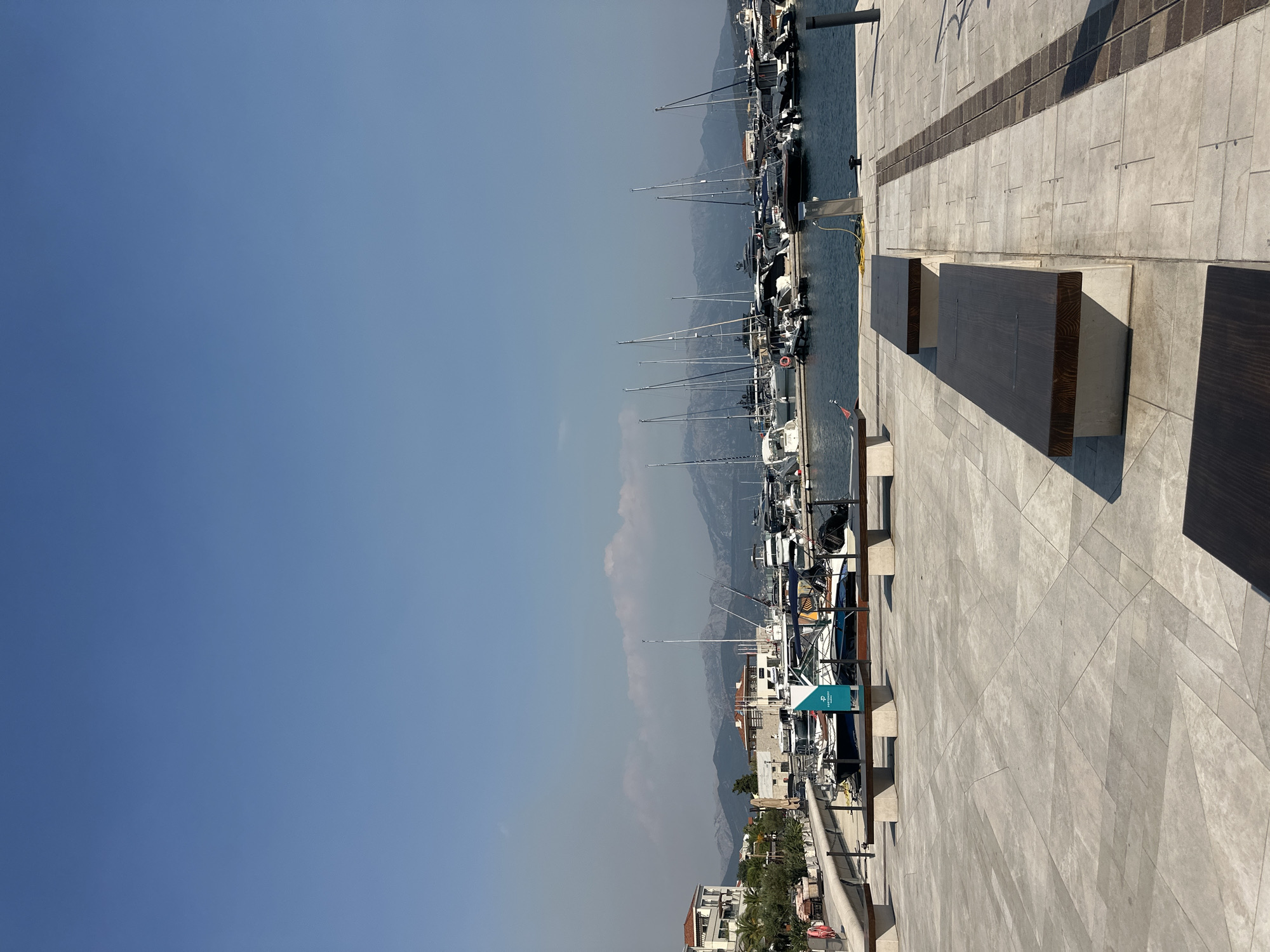 A wide-angle, eye-level shot captures a serene harbor scene in Kumbor, Montenegro, under a clear blue sky. In the foreground, a paved walkway with a geometric design features several dark wooden benches with concrete bases, some partially in shadow. The walkway leads to a calm, dark blue body of water where numerous boats are docked. These vessels range from sleek yachts to smaller motorboats and sailboats, their masts creating a dynamic silhouette against the backdrop.
Beyond the harbor, a waterfront promenade is lined with buildings, some with terracotta roofs and balconies, suggesting a residential or commercial area. Greenery, including trees and bushes, is visible along the promenade. In the distance, a range of majestic mountains forms a rugged horizon, their peaks softened by a slight haze. The overall atmosphere is one of peaceful, sunny leisure in a picturesque coastal town.