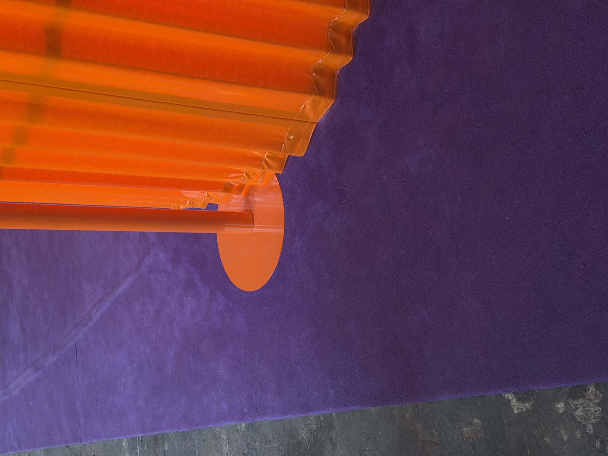 The image shows a section of a colorful display or art installation, likely located in Milan, Italy, given the context. An orange corrugated plastic structure is positioned against a purple textured wall or surface. The bottom of the image reveals a concrete floor. The plastic structure appears to be a series of parallel bars or panels covered in clear plastic, attached to a circular orange support. The purple surface occupies most of the image's background, while the concrete floor creates a bottom border. The overall impression is of a contemporary, brightly colored scene, potentially part of an art installation or a pop-up event in Milan. There is no evidence of people in this image.