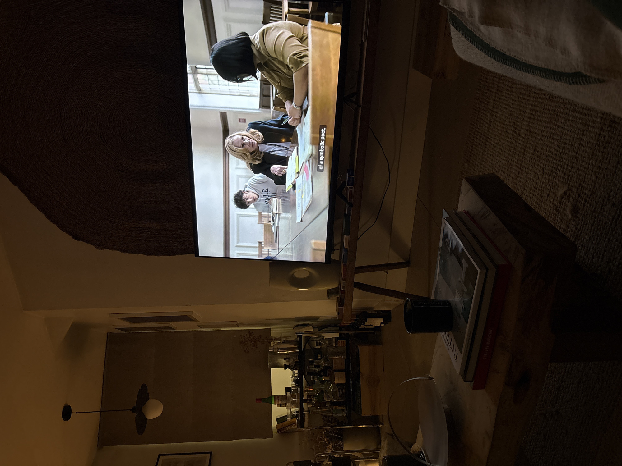 The image depicts a residential interior at night. A television displays a scene with three individuals gathered around a table in what appears to be a university or institutional setting. The characters are engaged in discussion and review of documents on the table. The text "of a symbolic poet" is visible as closed captioning. Below the TV, a bar cart is stocked with bottles and glassware. A ceiling fan and lighting fixtures are visible in the background. A stack of books is situated on a surface adjacent to the TV. The location is likely a private residence with a home entertainment setup. The specific location within London is not discernible from the provided image.
