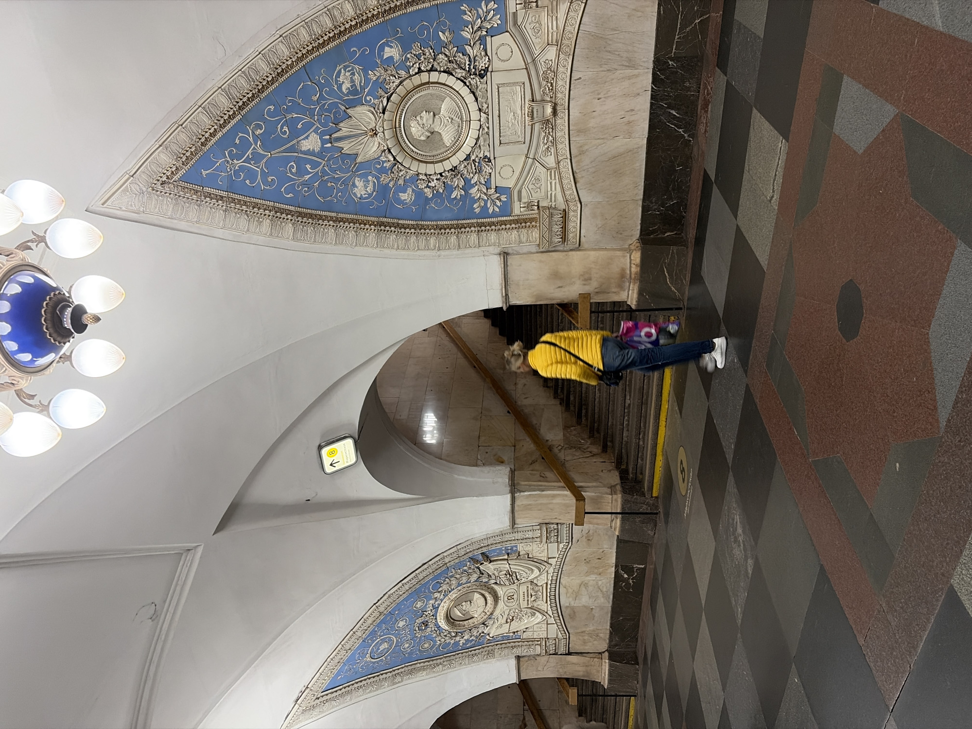 The image depicts an interior scene, likely a subway station, with ornate architecture. A woman wearing a yellow jacket and jeans is standing on the checkered floor, facing away from the camera and looking towards a staircase. She is carrying a shopping bag. The walls are decorated with blue panels featuring intricate white reliefs and portraits. A large, decorative chandelier hangs from the vaulted ceiling. The floor is paved with large square tiles in shades of black, grey, and red. An illuminated sign indicating "8" and an arrow points upwards, suggesting a pathway or platform number. The environment is clean and well-lit, with a marble-like finish on some architectural elements.