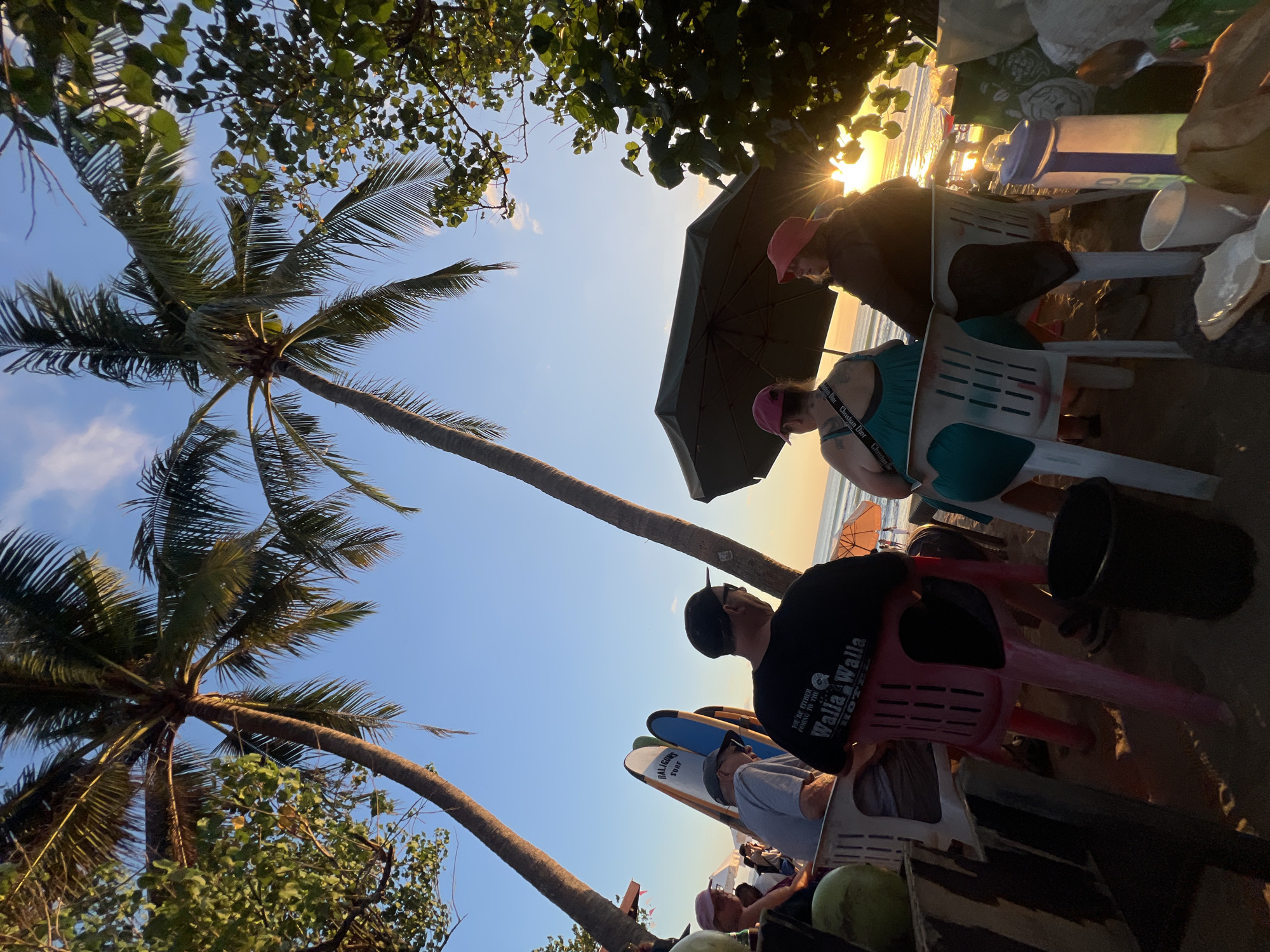 This image depicts a group of people seated at an outdoor beachside establishment in Kuta, Indonesia, during sunset. The foreground is dominated by several chairs, some empty and some occupied by individuals facing away from the camera, towards the ocean. A dark green umbrella provides shade for two women in the foreground who are wearing pink hats.
To the left, a man wearing a black t-shirt with a "Walla Walla Hotel" logo and a cap is visible. Beside him, another man wears a grey t-shirt and a cap. Behind them, several surfboards are propped up against a low structure.
The environment is a sandy beach with palm trees arching over the scene, their fronds silhouetted against a clear blue sky with a few wispy clouds. The setting sun casts a warm glow on the water, creating a serene and tropical atmosphere. Several plastic cups and a water jug are visible on a table in the foreground, suggesting a casual dining or relaxation setting. In the distance, more beachgoers and umbrellas are discernible, indicating a popular tourist spot.