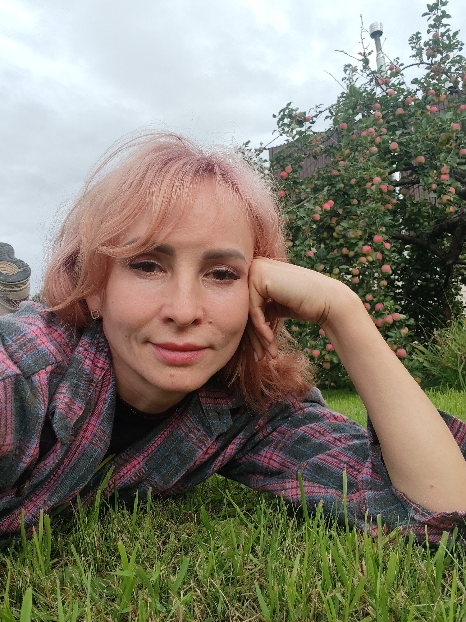 A woman with pink hair, wearing a pink and gray plaid shirt, lies on the grass. Her right arm is bent, and her head is resting on her hand. Behind her, an apple tree is laden with red apples. In the background, a wooden fence and a metal chimney are visible. The sky is overcast. The location is likely a backyard in Chaikovsky, Russia, given the presence of an apple tree and a wooden fence, typical features of Russian dachas (country houses).