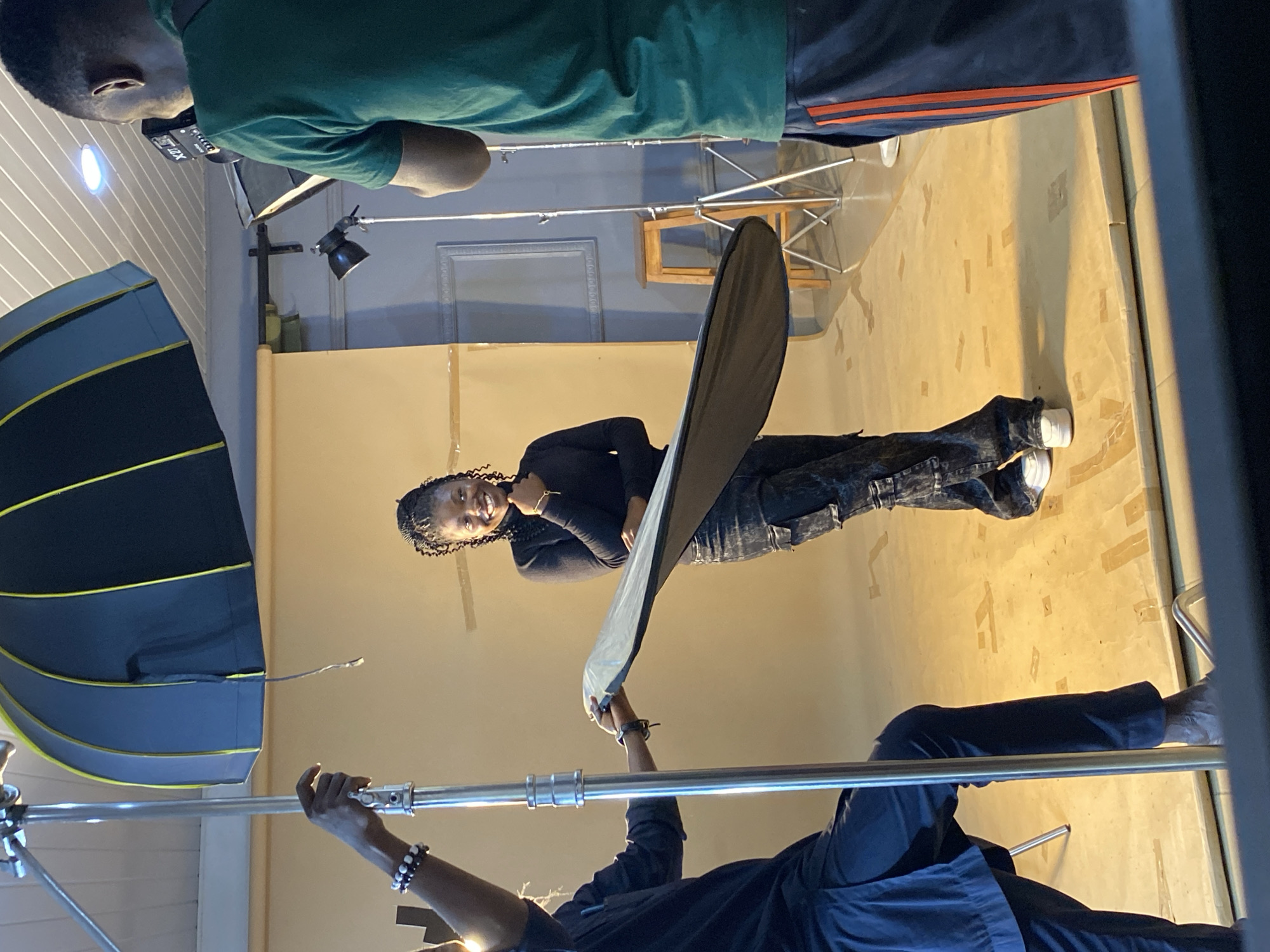 In a studio setting, a young Black woman with braided hair is posing in front of a tan backdrop. She is wearing a black turtleneck sweater and dark, distressed cargo pants with white sneakers. The woman is smiling and has her right hand gently touching her chin. Two individuals are assisting with the photoshoot. To the left, a person in a dark outfit is holding a silver light stand with a large, black and yellow softbox umbrella attached. This person is also holding a portable reflector. To the right, a person wearing a green t-shirt and dark shorts with orange stripes is holding a camera and appears to be taking pictures. The studio floor is a light tan color, with some tape markings visible. In the background, there is a wooden stool and a framed decorative piece on a blue wall. The overall scene depicts a professional photo session. The location is specified as Bukuru, Nigeria.