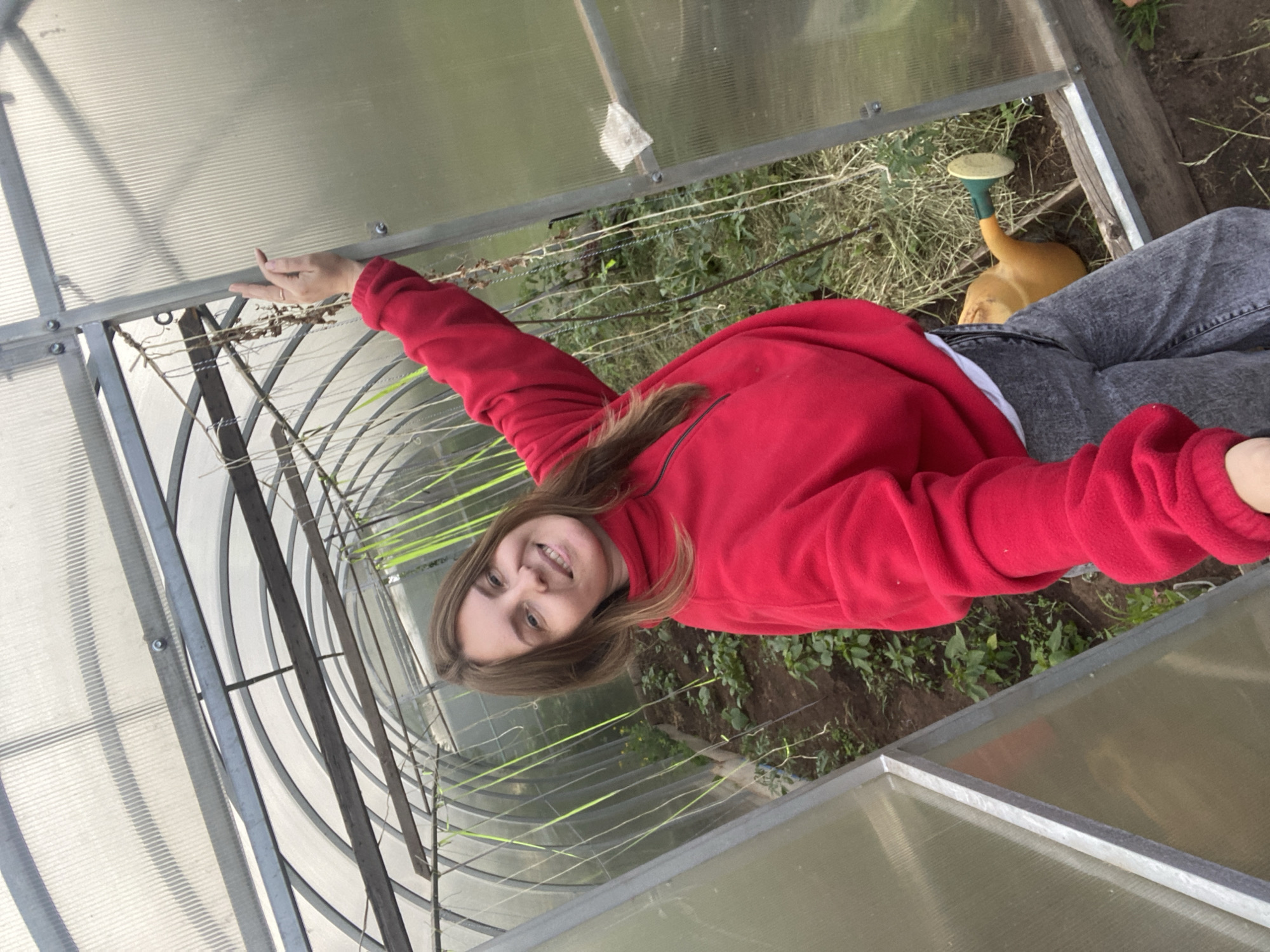 An adult female is captured in a selfie within a greenhouse, presumed to be located in Chaikovsky, Russia. She has light brown, shoulder-length hair and is smiling broadly, revealing teeth. She wears a red fleece half-zip pullover over a white undershirt and grey jeans. Her left hand, adorned with a thin ring on the ring finger, rests on the vertical metal frame of the translucent greenhouse wall to her right. The greenhouse features a corrugated translucent plastic or polycarbonate roof and walls supported by a curved metal frame. Internal wooden crossbeams are visible across the ceiling, from which multiple green and off-white strings or twine hang down, likely for supporting climbing plants. Plant beds with dark soil and small green foliage are visible on either side of a central dirt path, extending into the background. A yellow watering can with a green spout and cream-colored rose lies on its side on the ground in the bottom right corner. The lighting is bright and diffused.