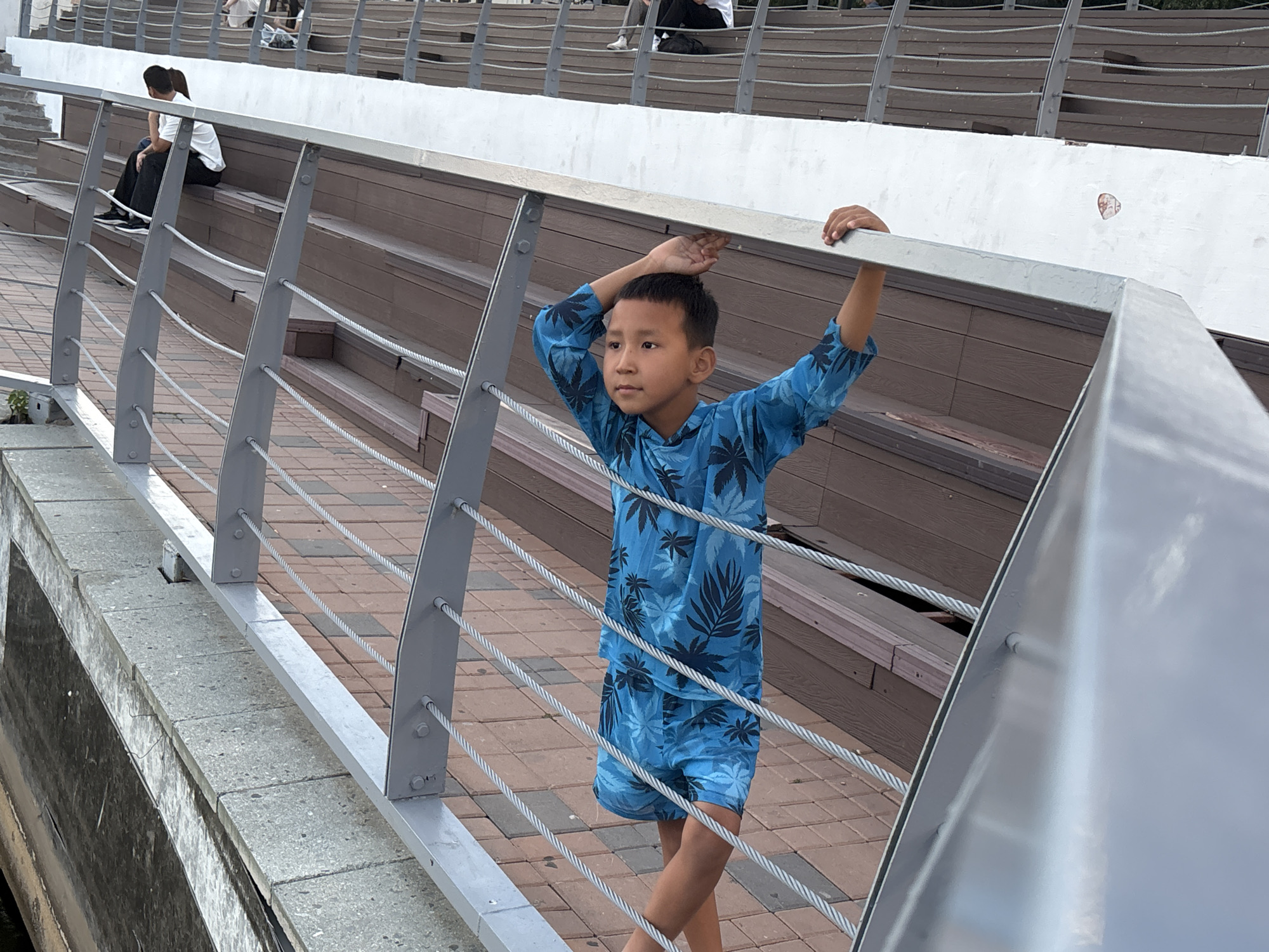 In a city park in Astana, Kazakhstan, a young boy stands by a railing, his hands resting on the metal bar above him. He is wearing a blue t-shirt and shorts with a leaf pattern. Behind him, people sit on wooden bleachers. The boy is looking to his left, his gaze directed away from the camera. The railing consists of metal posts connected by thin, braided steel cables. The bleachers are made of dark wood, and the ground is paved with reddish-brown bricks. The scene appears to be set during daytime, with overcast skies casting a diffused light.
