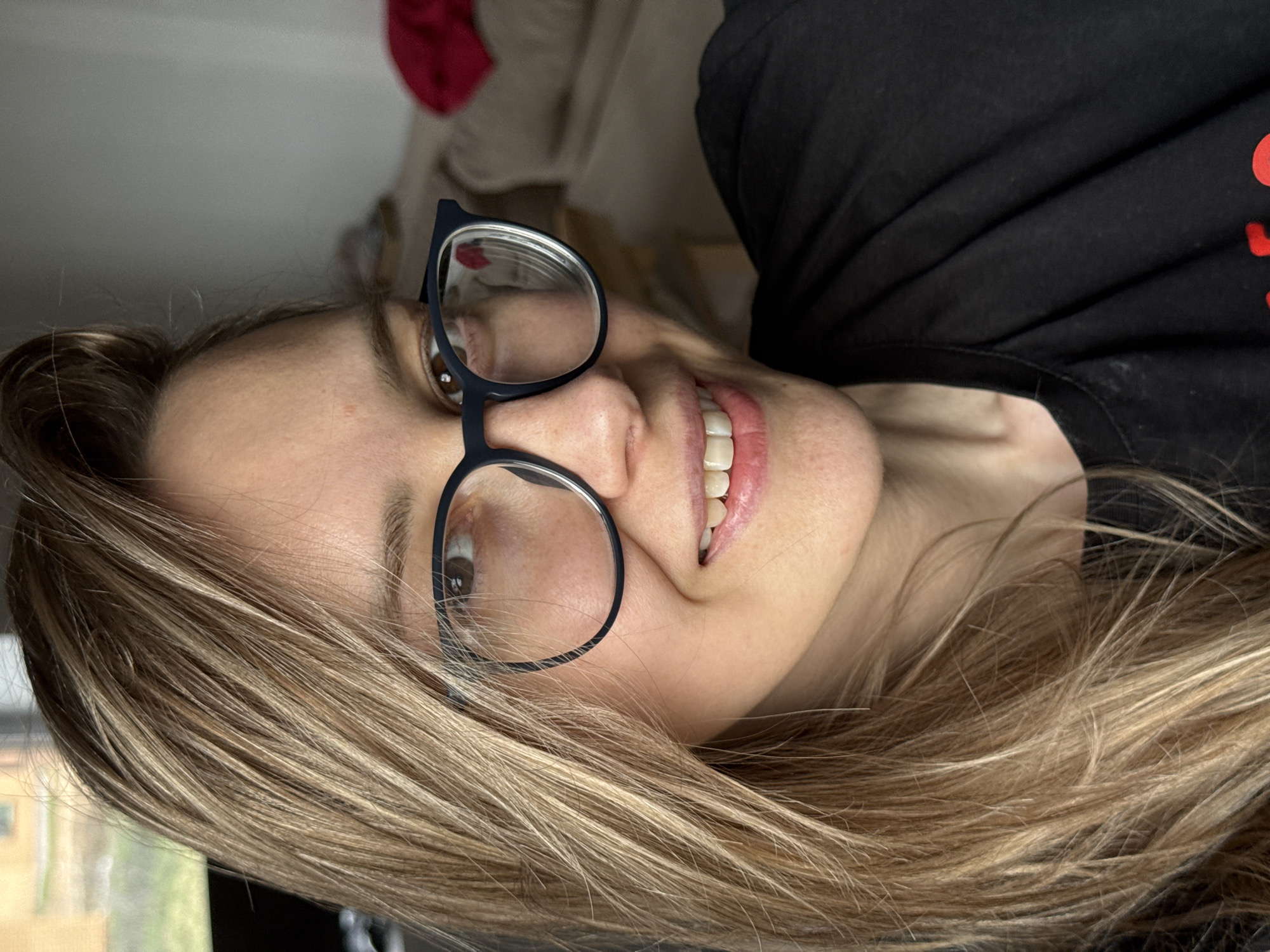 A woman with long blonde hair and wearing glasses smiles at the camera. She is wearing a black t-shirt. The background is blurred, but appears to be indoors. The location is unknown, and there is no evidence to suggest the city of Chaikovsky, Russia.
