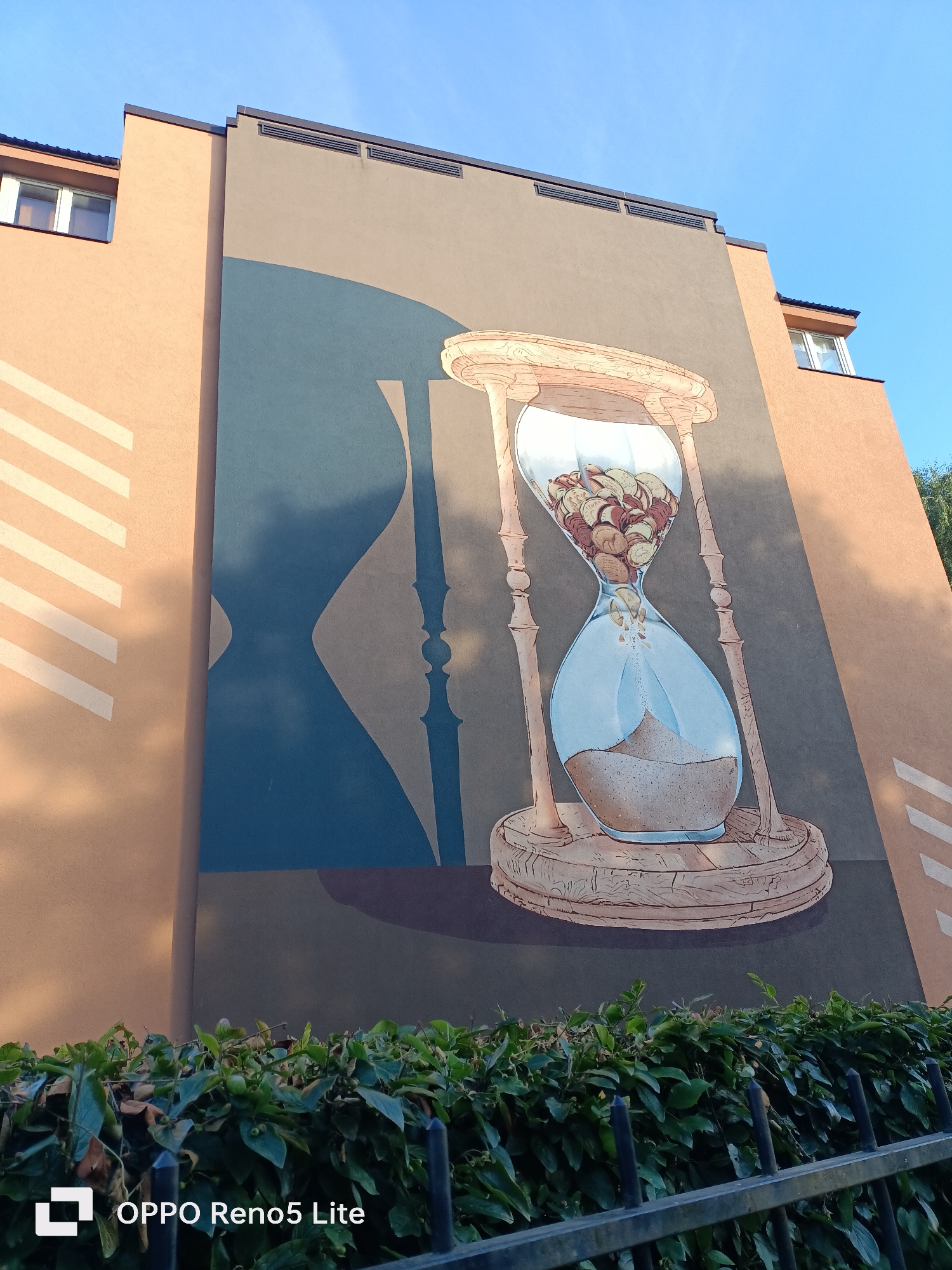 The image shows a mural of an hourglass on the side of a building. The hourglass is filled with coins, and sand is falling from the top bulb to the bottom. The building is painted in shades of beige and grey, with a large blue shadow on the wall. In the foreground, there is a hedge and a black metal fence. The sky is blue. The mural is located in Tallinn, Estonia.