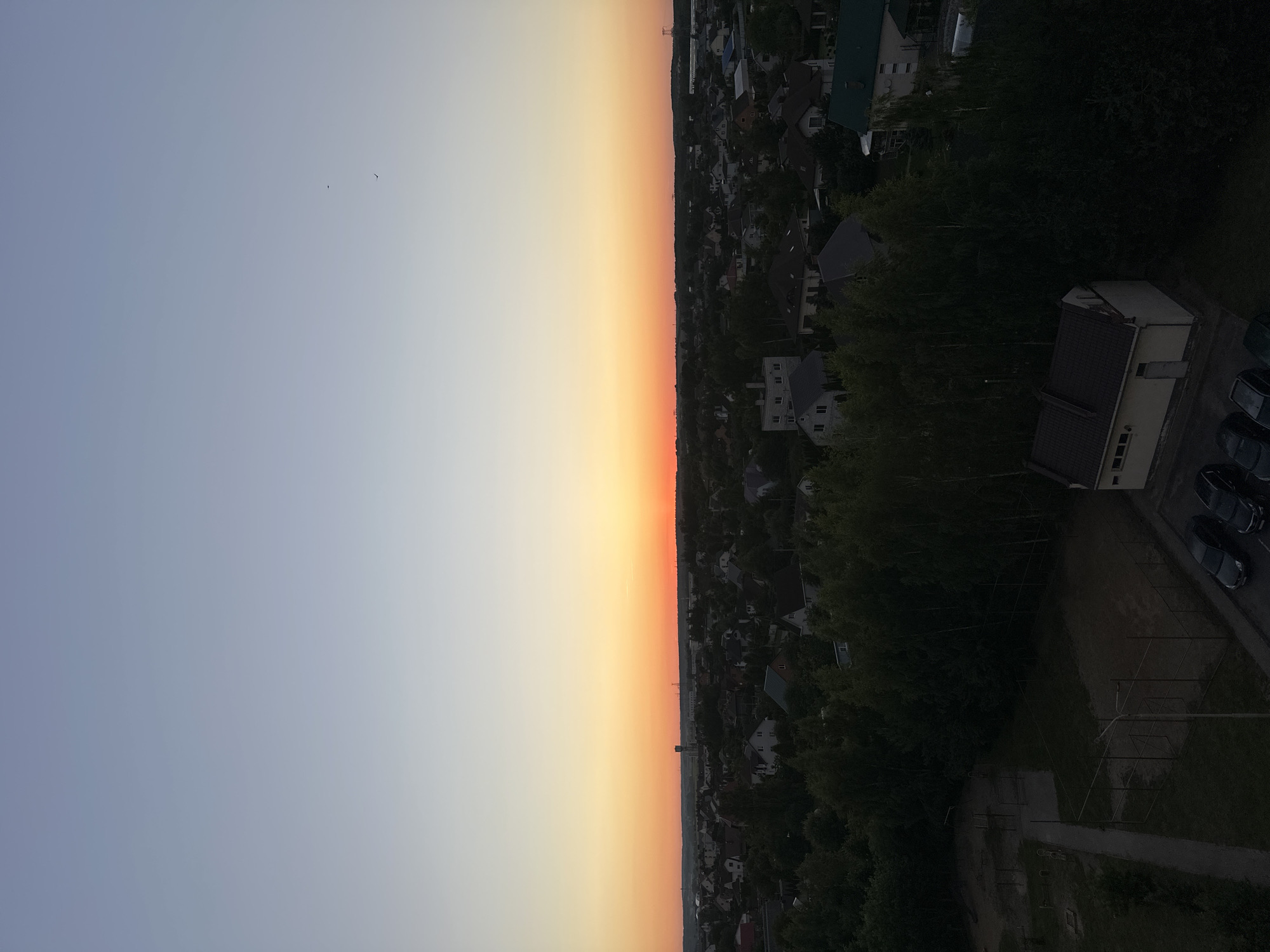 This image captures an aerial view of a residential area at dusk, likely in the vicinity of Шчомысліцкі сельскі Савет, Belarus. The sky transitions from a pale blue at the top to a vibrant orange and red horizon line, indicating the setting sun. Below the horizon, a dense expanse of trees dominates the foreground, interspersed with houses featuring various roof styles, some with visible chimneys. A few taller structures, possibly communication towers or industrial elements, are discernible in the distance. In the lower part of the frame, a small utility building with a dark roof is visible, adjacent to a paved area with several parked cars, all dark in color. A fenced recreational area, possibly a sports court or playground, with some equipment, is also present in the lower left portion of the image. Two small birds are visible in the upper portion of the sky. The overall scene depicts a quiet, semi-urban or suburban landscape as daylight fades.