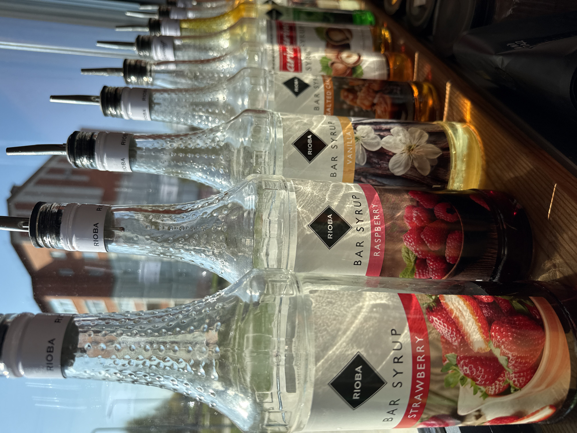 A row of Rioba brand bar syrup bottles sits on a shelf. The bottles are glass with a textured surface and white labels featuring a black diamond Rioba logo. The flavor of each syrup is indicated on the label with an image of the respective fruit or ingredient. From front to back, the flavors are Strawberry, Raspberry, Vanilla, Salted Caramel, and Hazelnut. Further back, other syrup bottles with lighter, golden-colored syrups can be seen. The bottles have metal pour spouts. A window reflects the outside; a multi-story brick building is visible. It is not possible to identify the city of Votkinsk from the image.