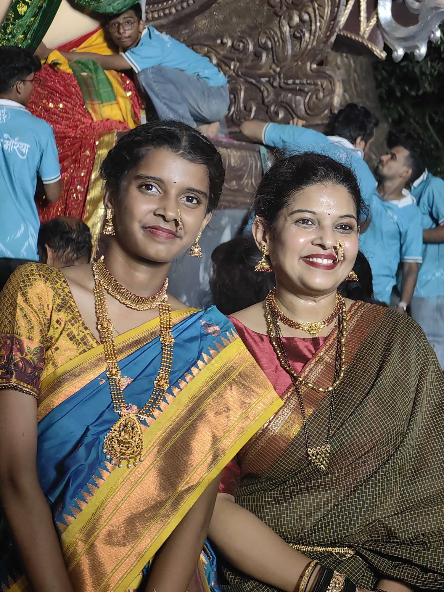 In Mumbai, India, two women are posing for a portrait. The woman on the left is wearing a blue and yellow sari with gold jewelry. The woman on the right is wearing a dark green and brown sari with gold jewelry and a black mangalsutra. Both women are smiling. Behind them, several men in blue t-shirts are visible, some are interacting with a decorative structure. The setting appears to be an outdoor event.