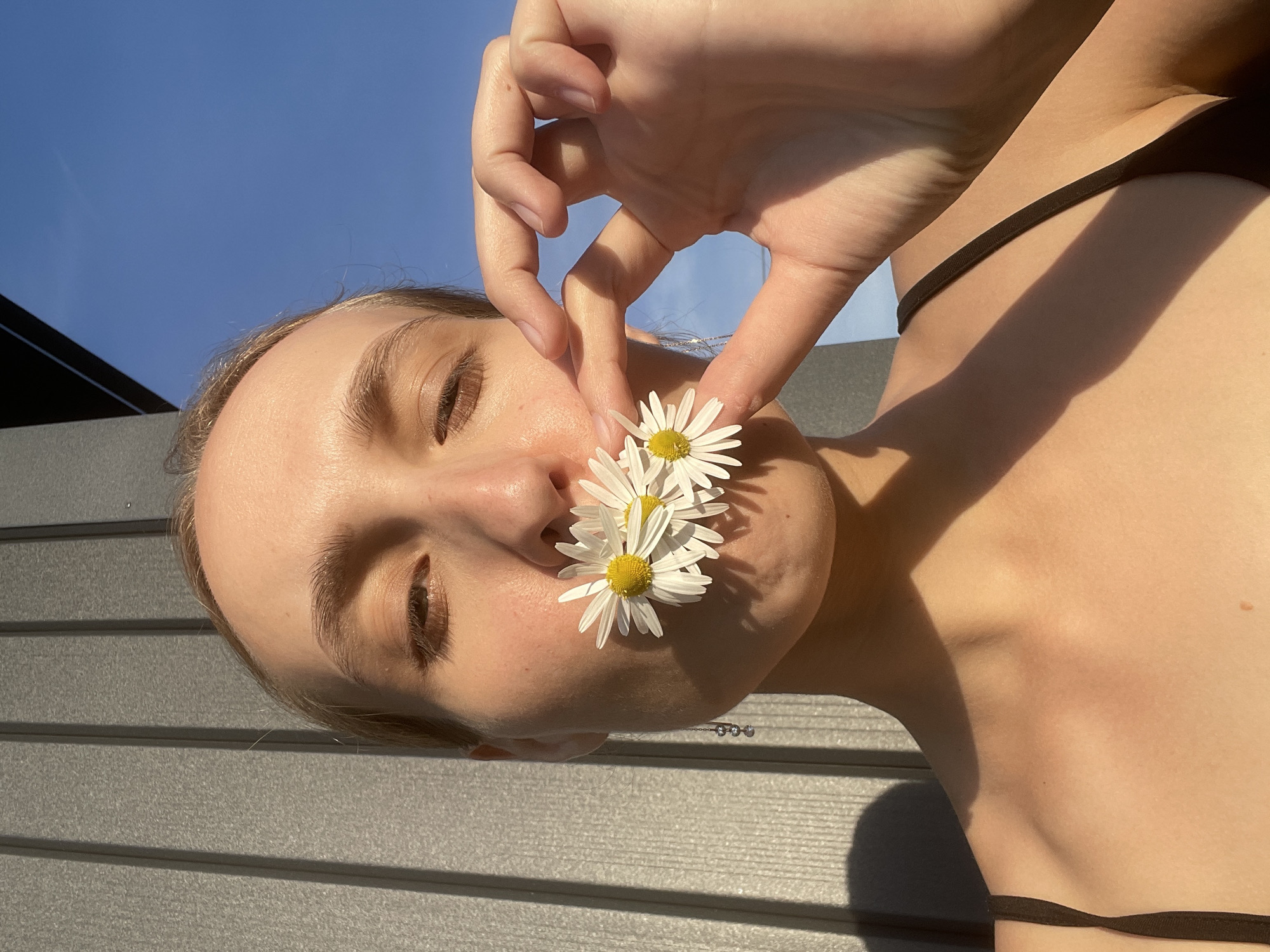 A young, light-skinned individual is holding three small daisies between their nose and index finger. The person has brown hair pulled back, closed eyes, and is wearing a black string top. The background shows a clear blue sky above a dark, low building and a gray, horizontal-planked surface, likely a deck or fence. The scene is bathed in sunlight, casting shadows across the person's face and body. The location in Подшивалово, Russia is not evident from the image.