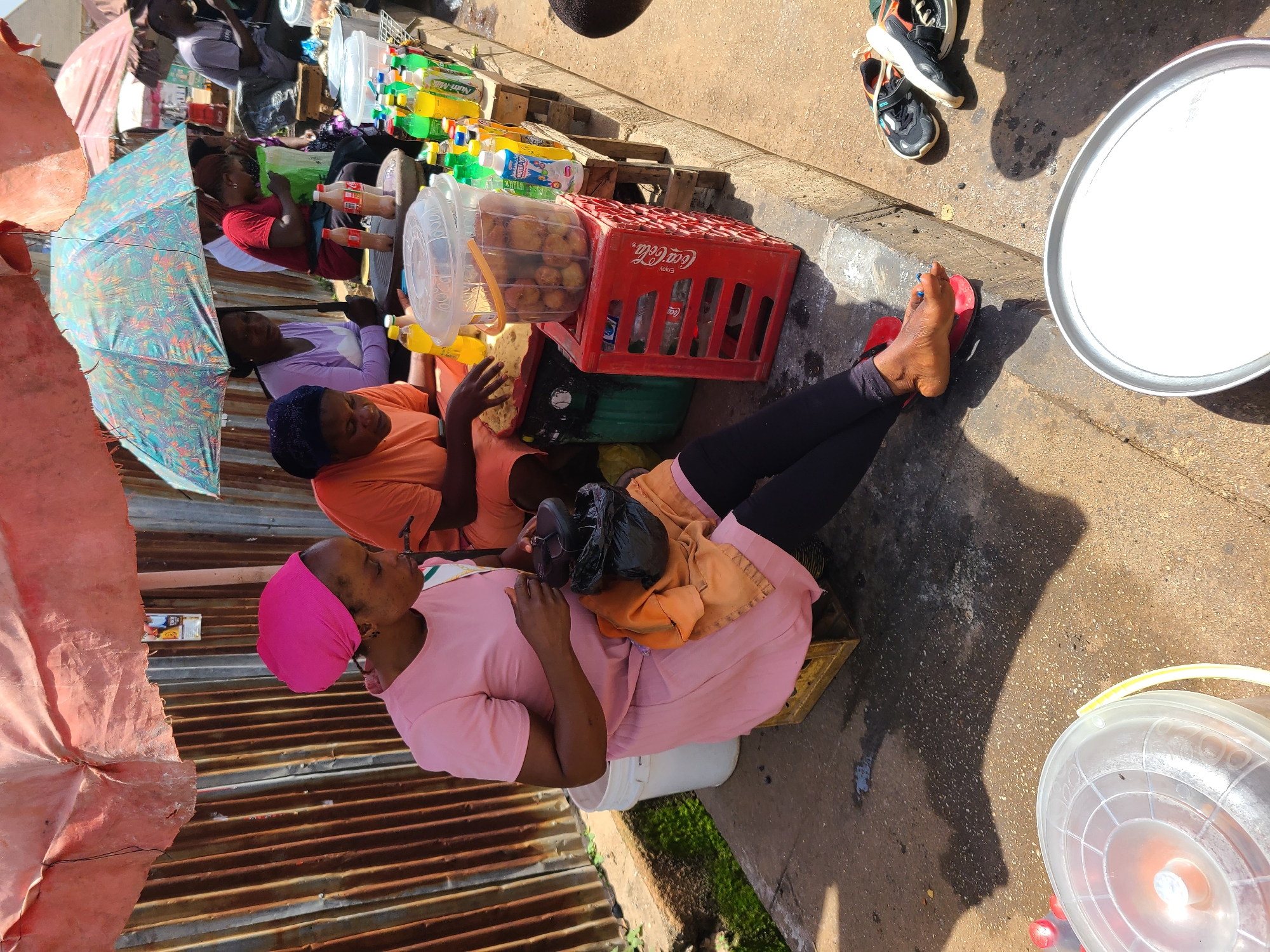 In Bukuru, Nigeria, a street vendor, a woman wearing a pink headscarf and a pink dress, sits on a wooden crate. She is preparing food, which is contained in a black plastic bag, and is covered with an orange cloth draped over her lap. To her right, another woman in an orange shirt is seated and also appears to be preparing food. They are both situated beneath a makeshift stall constructed with corrugated metal sheeting and covered by umbrellas. A variety of bottled drinks, including Coca-Cola, are displayed on wooden planks to the right of the women. A large red Coca-Cola crate is visible, holding a clear plastic container filled with fried food items. In the foreground, a large silver tray rests on the ground, and a white plastic bucket is positioned to the left of the seated vendor. The ground is paved with concrete, and shadows from the stall and people are cast across it. A pair of sneakers lies on the ground near the curb.