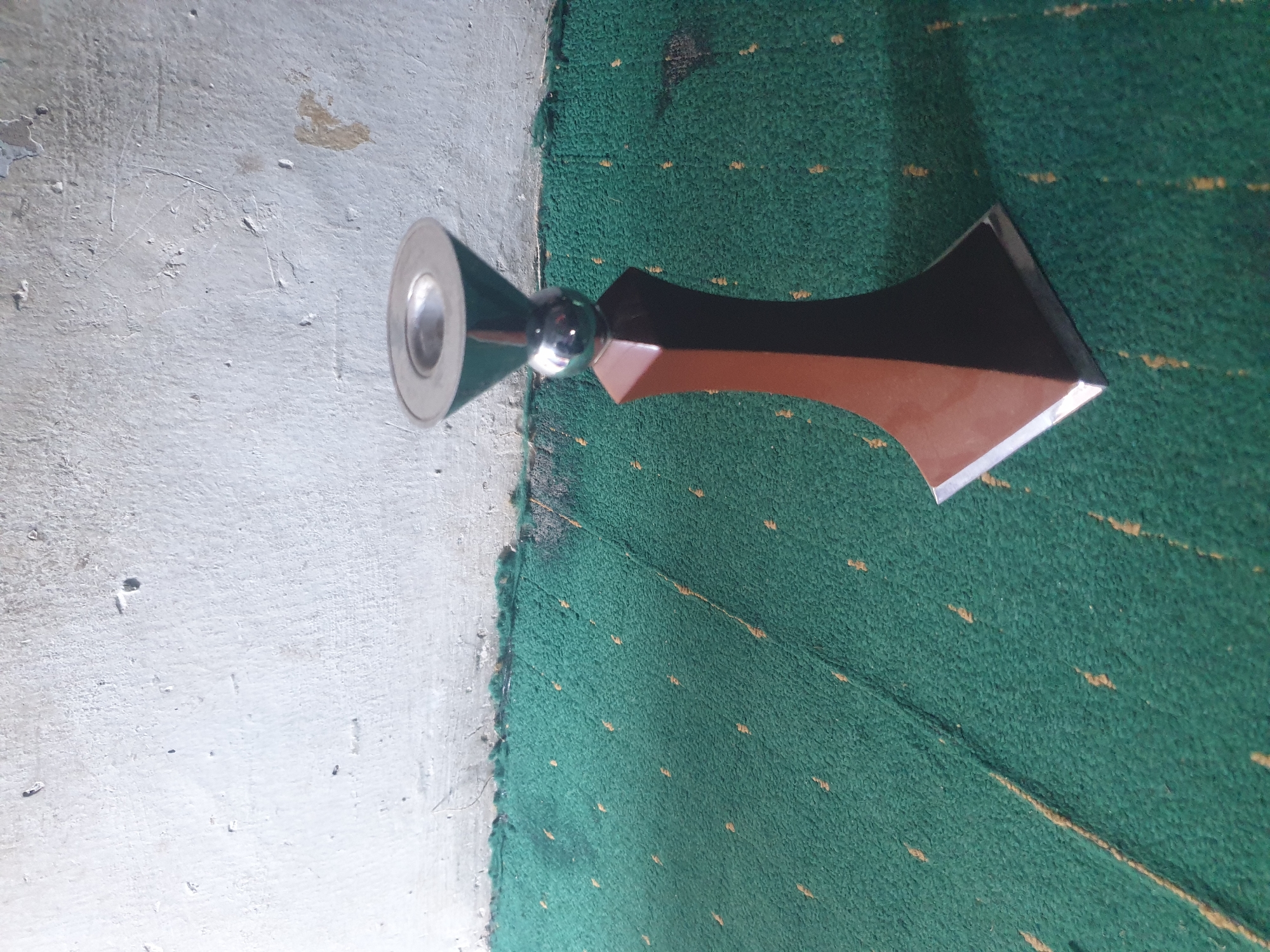 In the city of Unknown, Unknown, a vertical image captures a decorative object positioned on a green carpet against a light-colored wall. The object, approximately 30-40 cm in height, features a multi-material and multi-component construction. Its uppermost section is a silver-colored, metallic, circular disk with a central depression. This sits atop a polished, spherical, silver-colored metallic element. Below the sphere is a tapered, multi-faceted (likely hexagonal or octagonal) body made of a dark brown, possibly wooden or composite, material. The base of the object is a square, silver-colored, metallic plate. A distinct shadow from the object extends to its right across the carpet.
The ground surface consists of a green carpet, characterized by a textured, short-pile weave and numerous small, irregular, light-brown/yellowish flecks distributed across its surface. Visible linear indentations or creases are present on the carpet. The carpet terminates against a wall that occupies the upper portion of the frame. The wall is light grey or off-white, exhibiting a rough, textured surface consistent with aged plaster or concrete. Multiple imperfections are visible on the wall, including small chips, minor cracks, and areas of discoloration or paint erosion, particularly towards the bottom and upper right. The interface between the carpet and the wall is uneven and shows signs of accumulated dust or debris. No human figures or discernible actions are present within the scene.