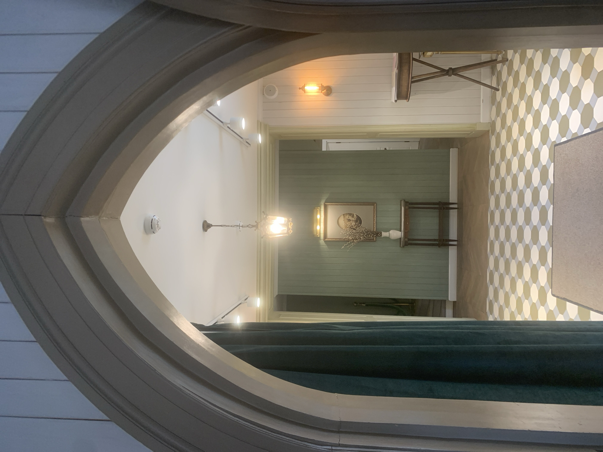 A view through an arched doorway, centered on a hallway. The hallway features green, horizontally paneled walls and a painting in a gold frame with a white vase on a table beneath it. A chandelier hangs from the white ceiling. A doorway is visible at the end of the hallway. The foreground features dark green fabric that is likely a curtain. The background includes a patterned floor with beige and white hexagons. The location is most likely in Arkhangel’skoye, Russia, given the prompt's specification.