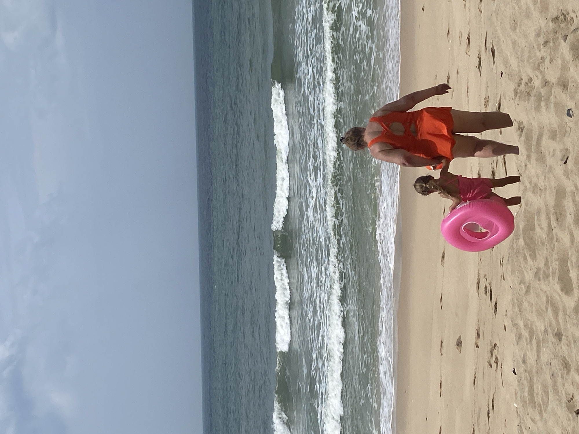 In Mũi Né, Vietnam, two individuals, an adult and a child, stand on a sandy beach facing the ocean. The adult, wearing an orange one-piece swimsuit with a distinctive cut-out back detail, holds their left hand out, seemingly guiding the child. The child, dressed in a pink swimsuit, carries a bright pink inflatable ring in their left hand. Gentle waves lap the shore, creating a serene beach scene. The sky is overcast, with muted tones suggesting a cloudy day. The ocean stretches out to the horizon, a deep blue-green. The sand is a light brown, marked with footprints, indicating recent activity on the beach. The overall environment suggests a relaxed, perhaps overcast, day at the seaside.