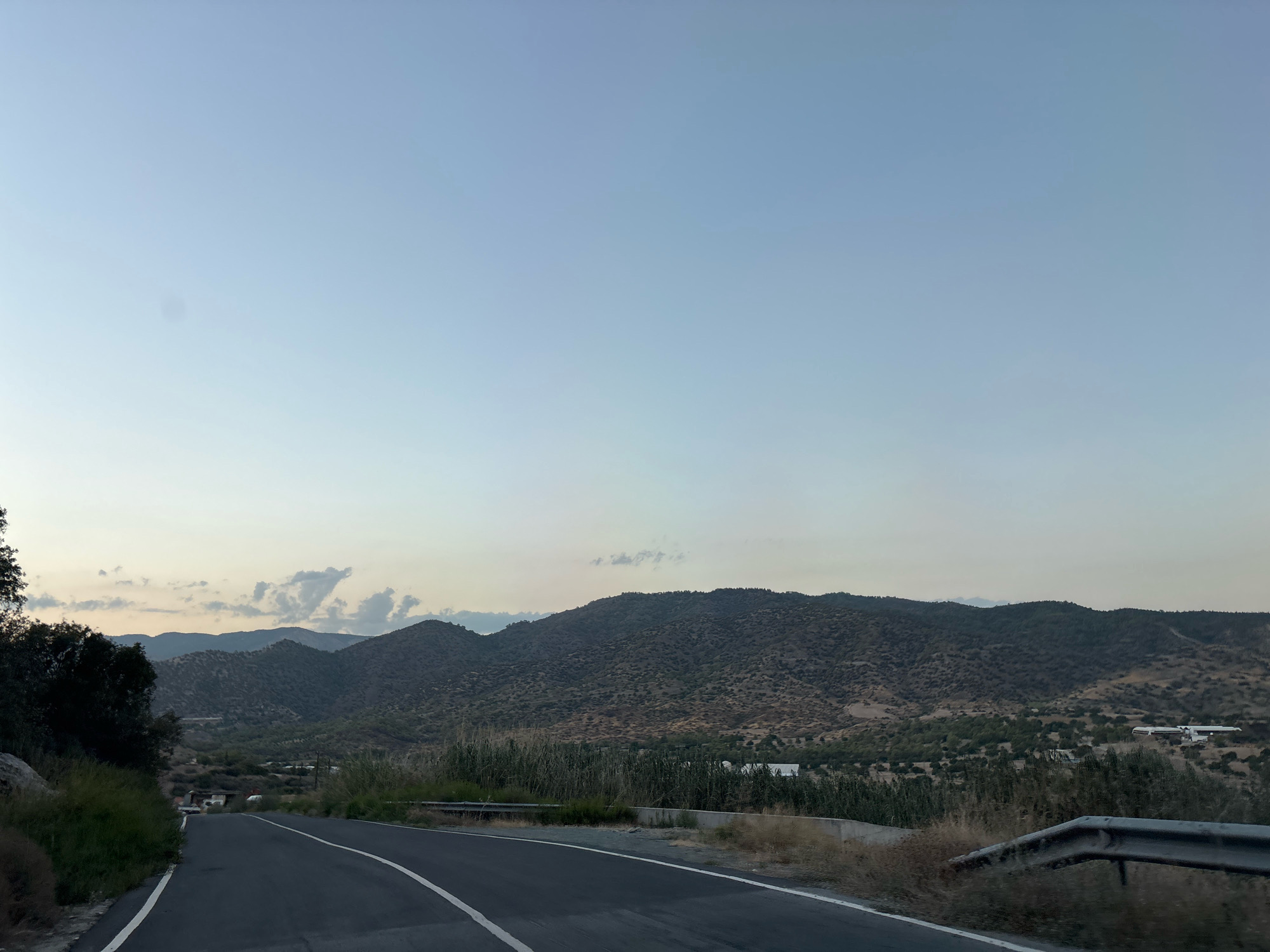 The image shows a road in a rural, mountainous area. The road curves to the left and is paved with asphalt, marked with white lines. On the left side of the road, there is dense vegetation and a large rock. On the right side, there is a metal guardrail and dry grass. In the background, there are rolling hills covered with trees and shrubs, with some buildings visible in the distance. The sky is a clear, pale blue, with a few wispy clouds. The overall impression is one of a natural landscape at dusk or dawn.