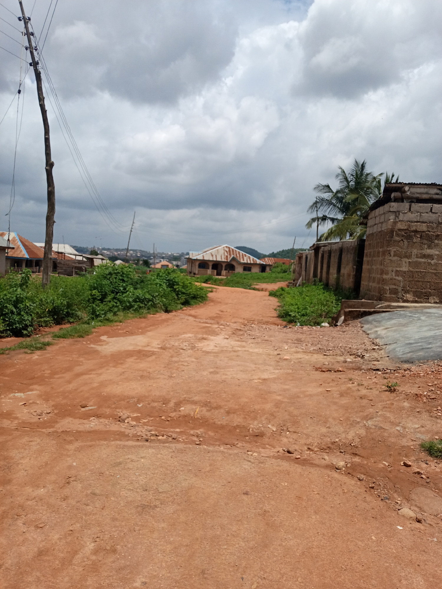 The image depicts a dirt road in Iseyin, Nigeria, under a cloudy sky. On the left side of the road, there is a wooden utility pole with electrical wires. The road is flanked by dense green vegetation and several buildings with corrugated metal roofs, some of which appear to be houses. To the right, there is a wall constructed from concrete blocks and more vegetation. In the distance, more buildings and hilly terrain can be seen. The overall scene suggests a rural or semi-urban setting within Iseyin.
