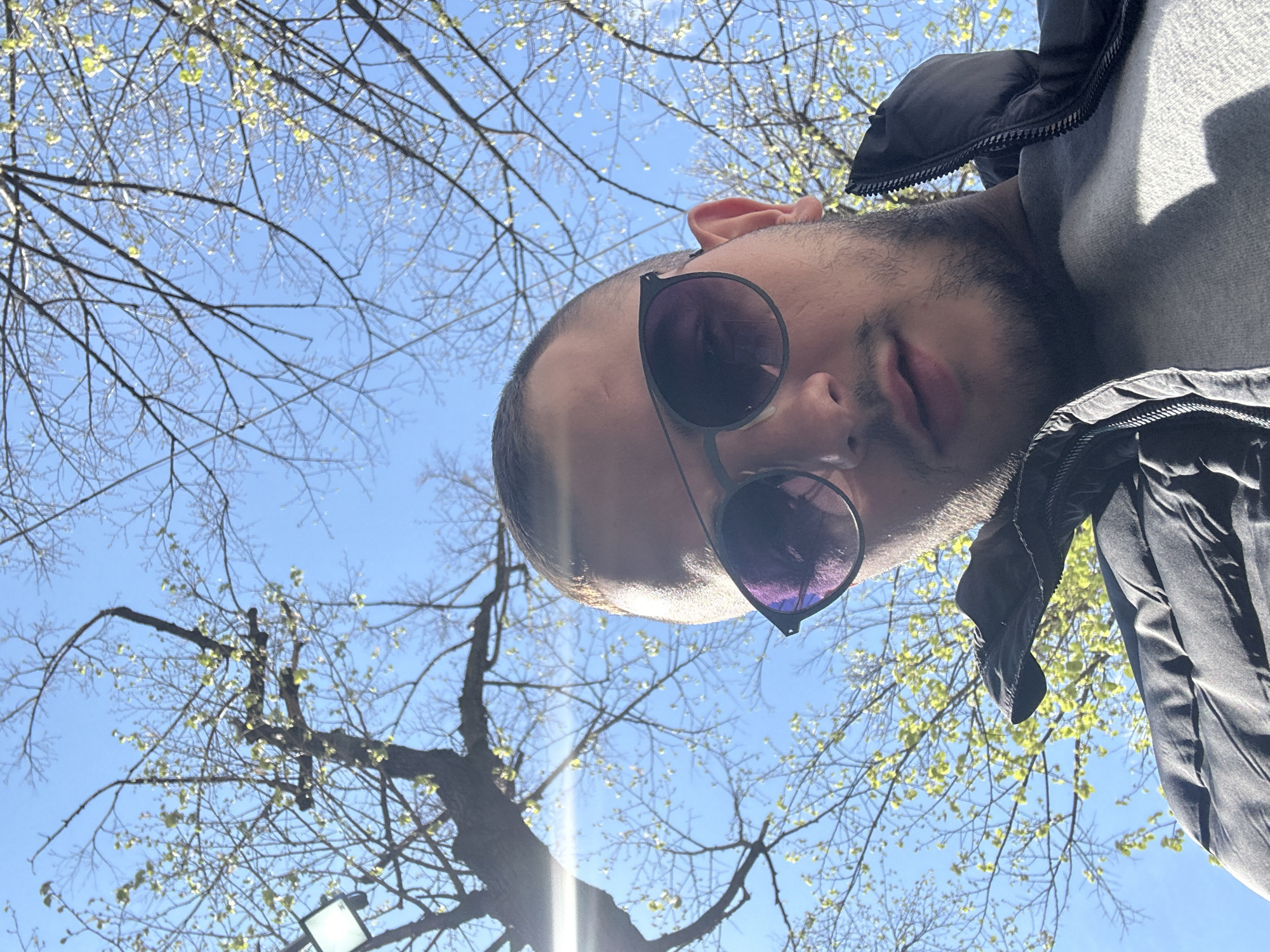 The image is taken from a low angle, looking up at a person wearing sunglasses and a jacket. Bare tree branches with emerging leaves fill the background, with a clear blue sky visible between them. There's a bright light source in the bottom left corner. The location is potentially a park or green space in Plovdiv, Bulgaria, considering the trees and typical urban environment.
