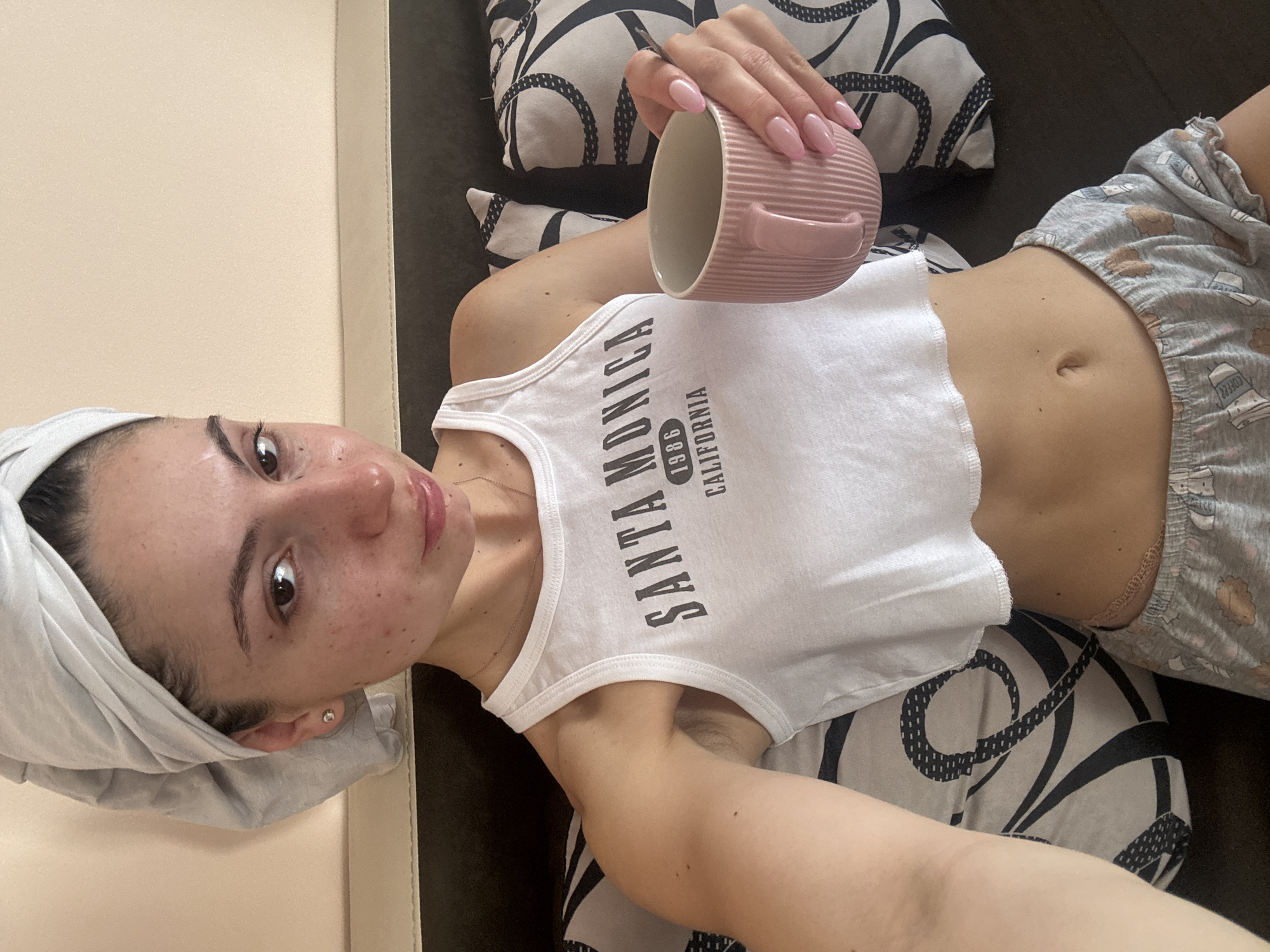 A young woman is captured in a relaxed, casual pose, lying down indoors, rotated 90 degrees counter-clockwise in the frame. Her head is wrapped in a light-colored towel, suggesting she might have just showered or is in the midst of a beauty routine. She has a natural complexion with some visible blemishes or freckles and glossy lips. A tiny stud earring is visible in her right ear.
She is wearing a white sleeveless crop top that reads "SANTA MONICA 1986 CALIFORNIA" in black letters. Her lower body is clad in light gray patterned shorts featuring small, cartoon-like graphics, possibly including coffee cups or animal shapes. A thin gold chain necklace can be seen on her chest.
In her right hand, she holds a light pink, ribbed ceramic mug with a small handle. Her fingernails are painted a soft pink, perfectly manicured. She is supported by two pillows with a distinctive black and white swirling or geometric pattern. A light-colored, possibly beige or cream, padded surface is visible on her left, suggesting she is on a bed or sofa.
The woman is looking directly at the camera with a calm, neutral expression. The scene suggests a quiet, personal moment of leisure or self-care within a domestic setting in Varna, Bulgaria. The lighting appears soft and even, typical of indoor environments.