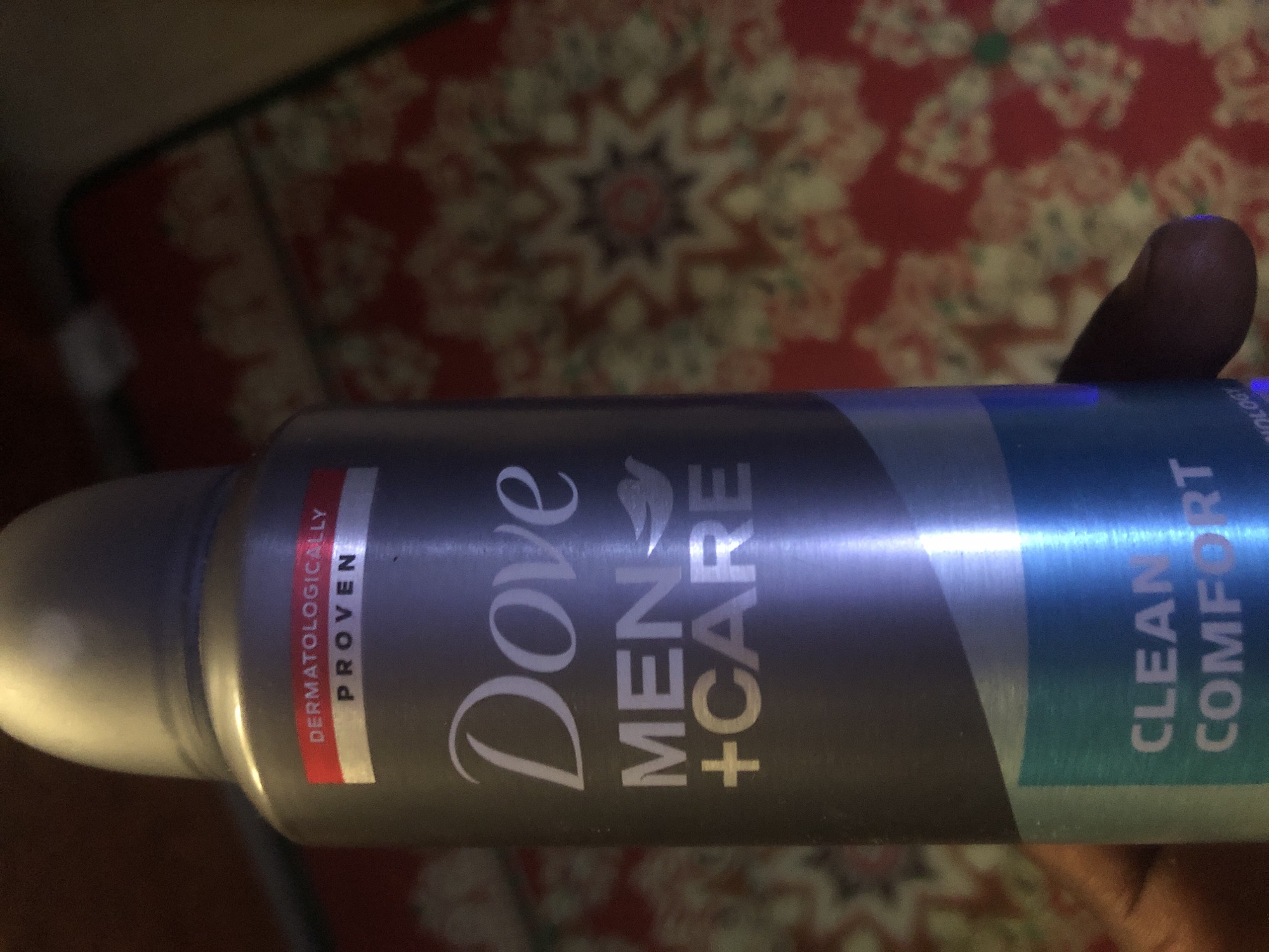 The image shows a close-up of a Dove Men + Care deodorant can, held by a person's hand which is partially visible in the lower right corner. The can is primarily grey with white and light blue lettering. A red and white label near the top reads "DERMATOLOGICALLY PROVEN". The brand name "Dove" is prominently displayed in white script, followed by "MEN + CARE" in capital letters. The bottom section of the can, in light blue, has text that appears to read "CLEAN COMFORT". The background is out of focus and shows a red fabric with a yellow floral pattern, possibly a rug or tapestry. The lighting suggests an indoor setting. There is no indication of the city of Kabong, Nigeria, or any urban environment in the image.