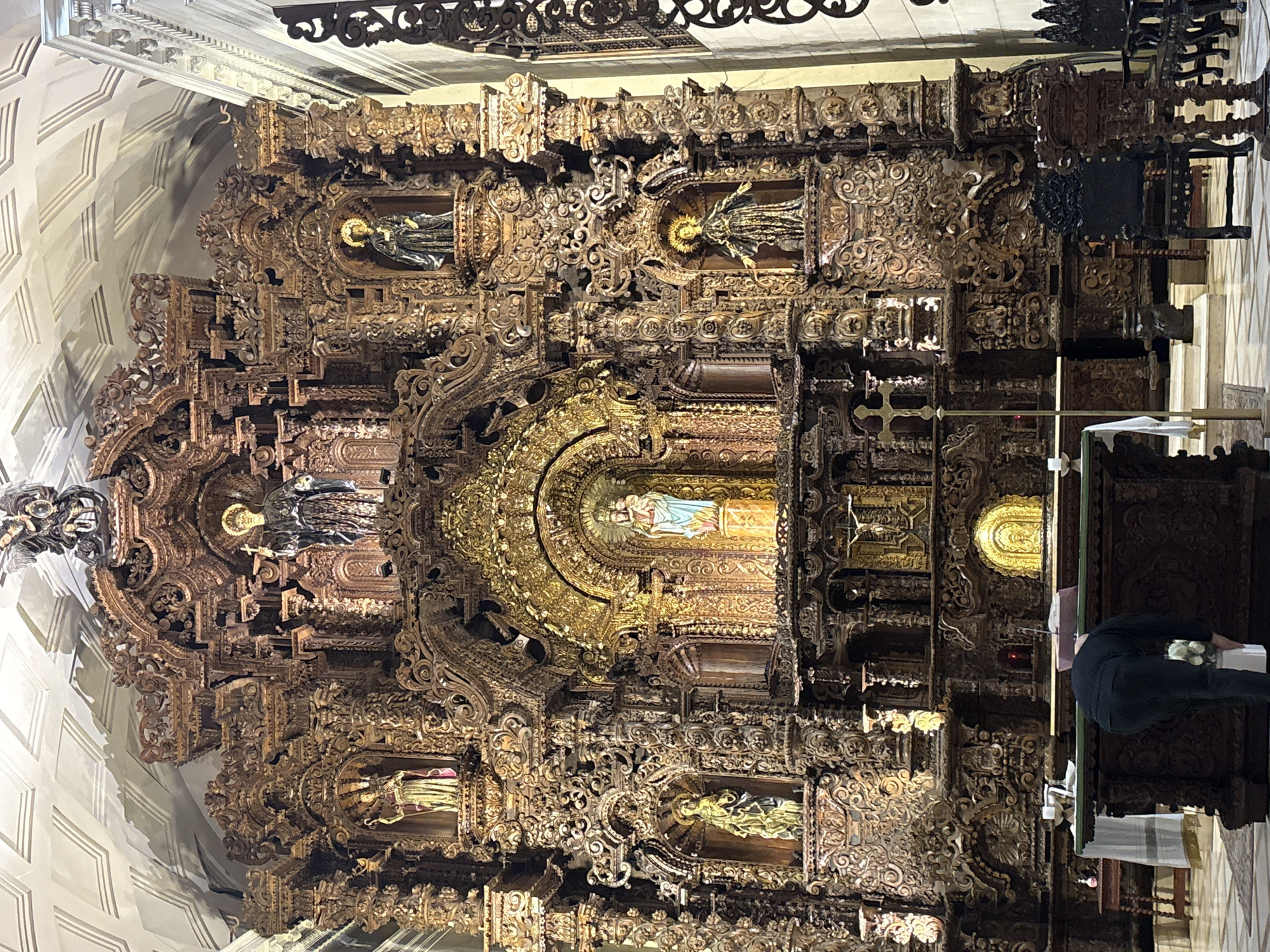 The image depicts the ornate interior of the San Isidro Cathedral in Lima, Peru. Dominating the scene is a large, intricately carved wooden altarpiece, likely made of gold-leafed cedar, adorned with multiple statues of religious figures, including the Virgin Mary and child at the center, illuminated by warm lights. In the foreground, a person in dark clothing is bending over a wooden altar, seemingly preparing for a service. The cathedral's interior features high ceilings with decorative plasterwork and other architectural elements typical of Baroque or Neoclassical styles. The flooring is a checkered pattern of light and dark tiles. The overall impression is one of grandeur and religious solemnity.