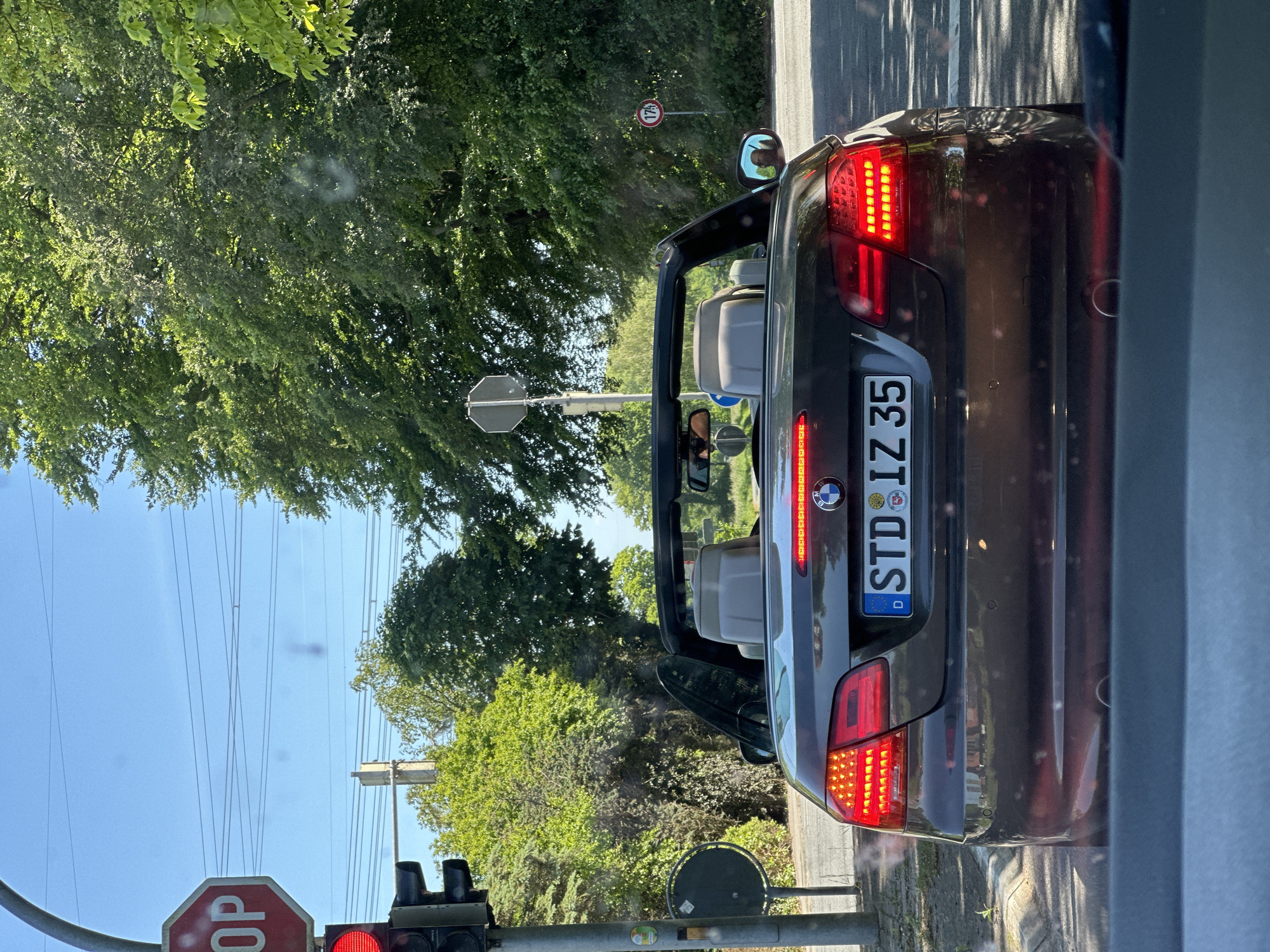 A medium, eye-level shot, looking straight ahead while slightly angled down, shows the rear of a dark convertible car with German license plate STD IZ 35. The convertible has an open roof. The scene is an urban or suburban setting with abundant greenery. The car is stopped at a traffic light, which is red. A stop sign is visible in the lower-left corner. The location is identified as Kutenholz, Germany, based on the license plate. A speed limit sign of 175 km/h is visible to the right of the car. Utility poles and overhead wires are visible against a blue sky in the background. A figure is partially visible in the rearview mirror of the convertible.