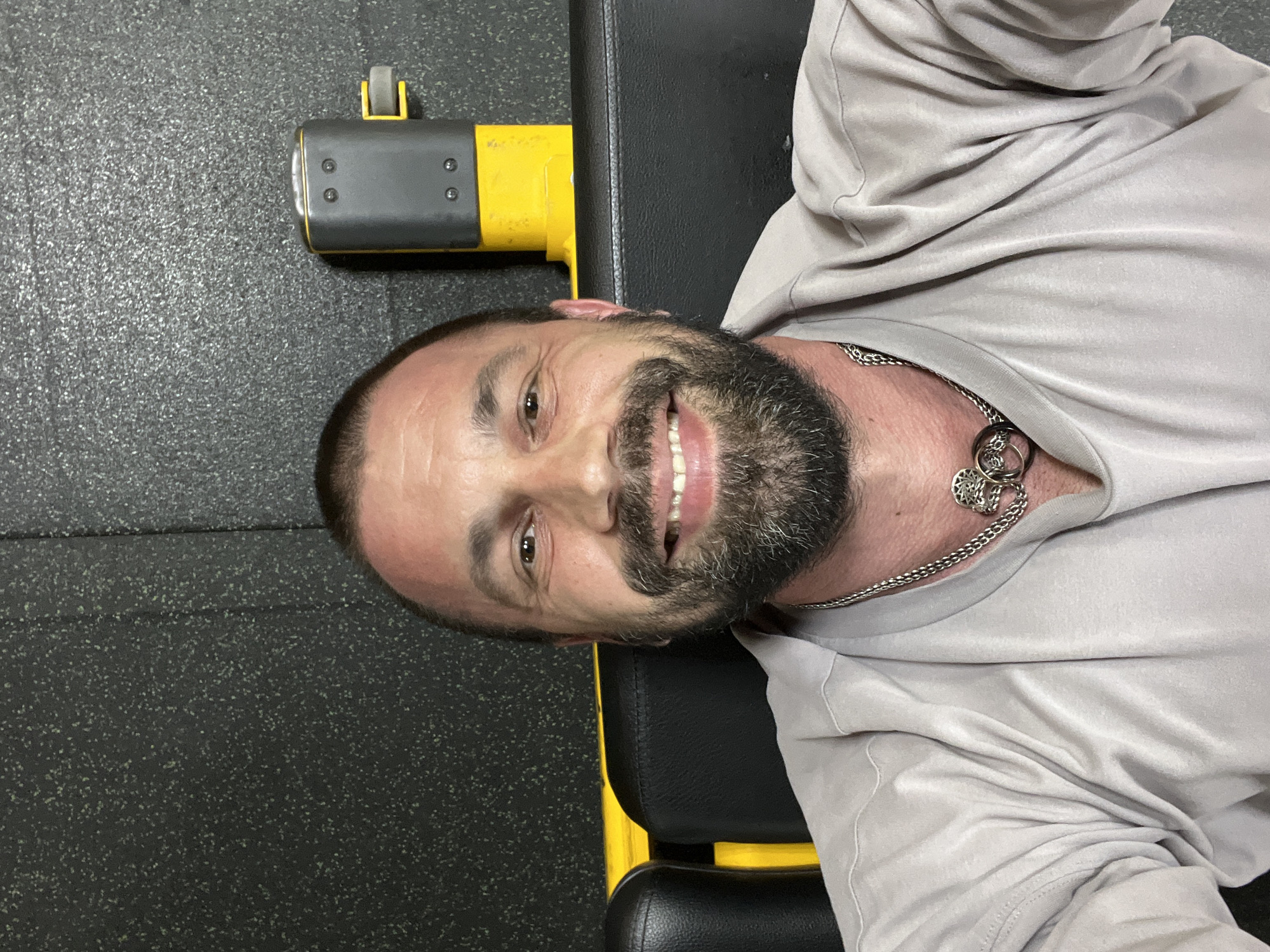 A man with a beard and short hair is taking a selfie in a gym. He is wearing a light-colored t-shirt and a silver necklace. Behind him, there is a black padded bench with yellow accents and a black textured wall. The scene is dimly lit. The image does not contain any information that would allow for its location in Yoshkar-Ola, Russia, to be determined.