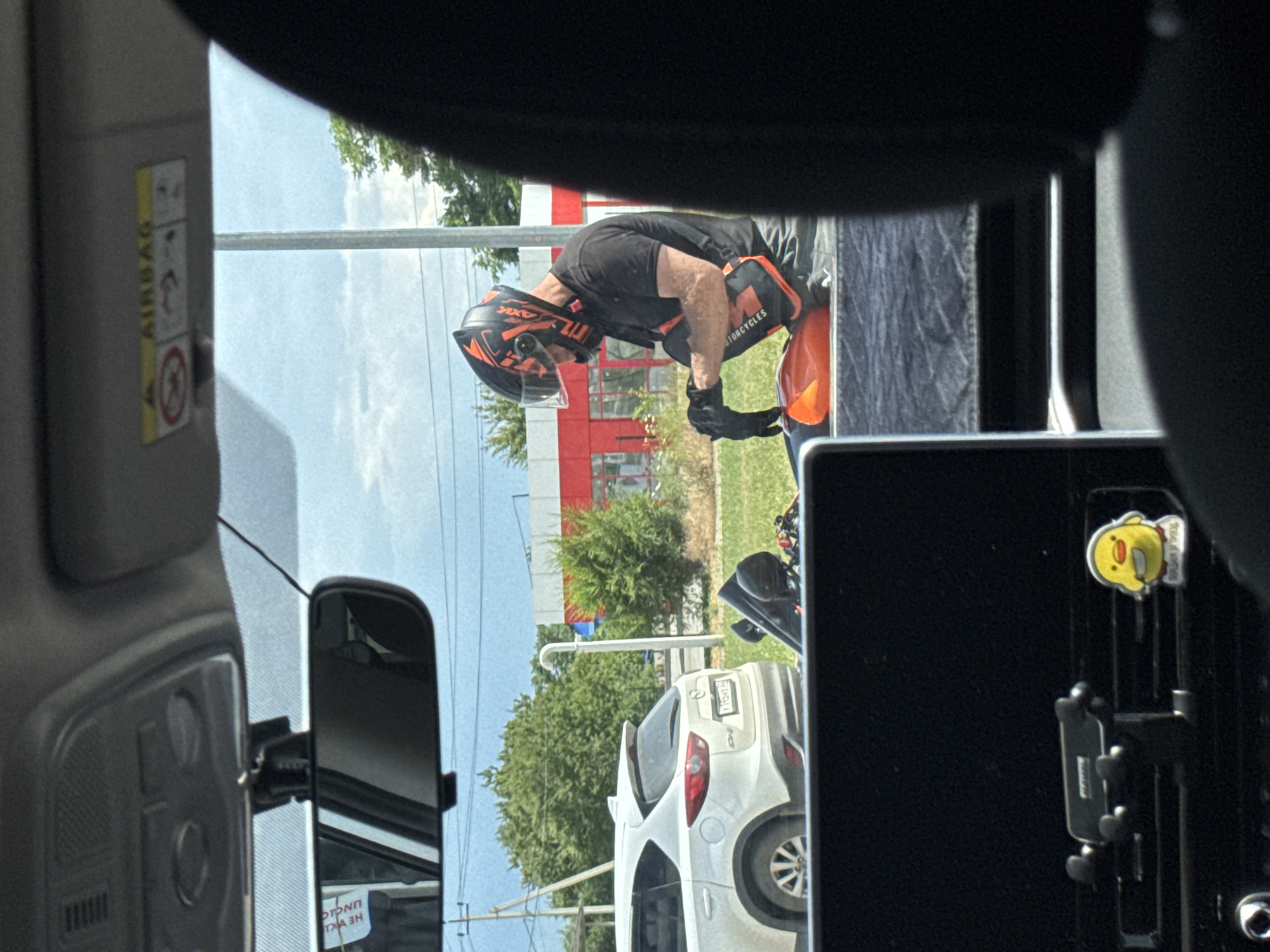 The image, captured from inside a vehicle, shows a man in a black t-shirt, orange and black motorcycle helmet, and orange motorcycle pants putting on black gloves. He is kneeling on what appears to be the seat of an orange motorcycle. The motorcycle is parked on a grassy area beside a road. In the background, a white SUV is parked. A building with red and white accents is visible behind the SUV, and beyond that, trees and a utility pole. The sky is cloudy. The license plate of the white SUV appears to be in Cyrillic script. The interior of the vehicle shows a rearview mirror, an airbag warning label, and a phone mount with a small yellow cartoon character attached to the air vent.