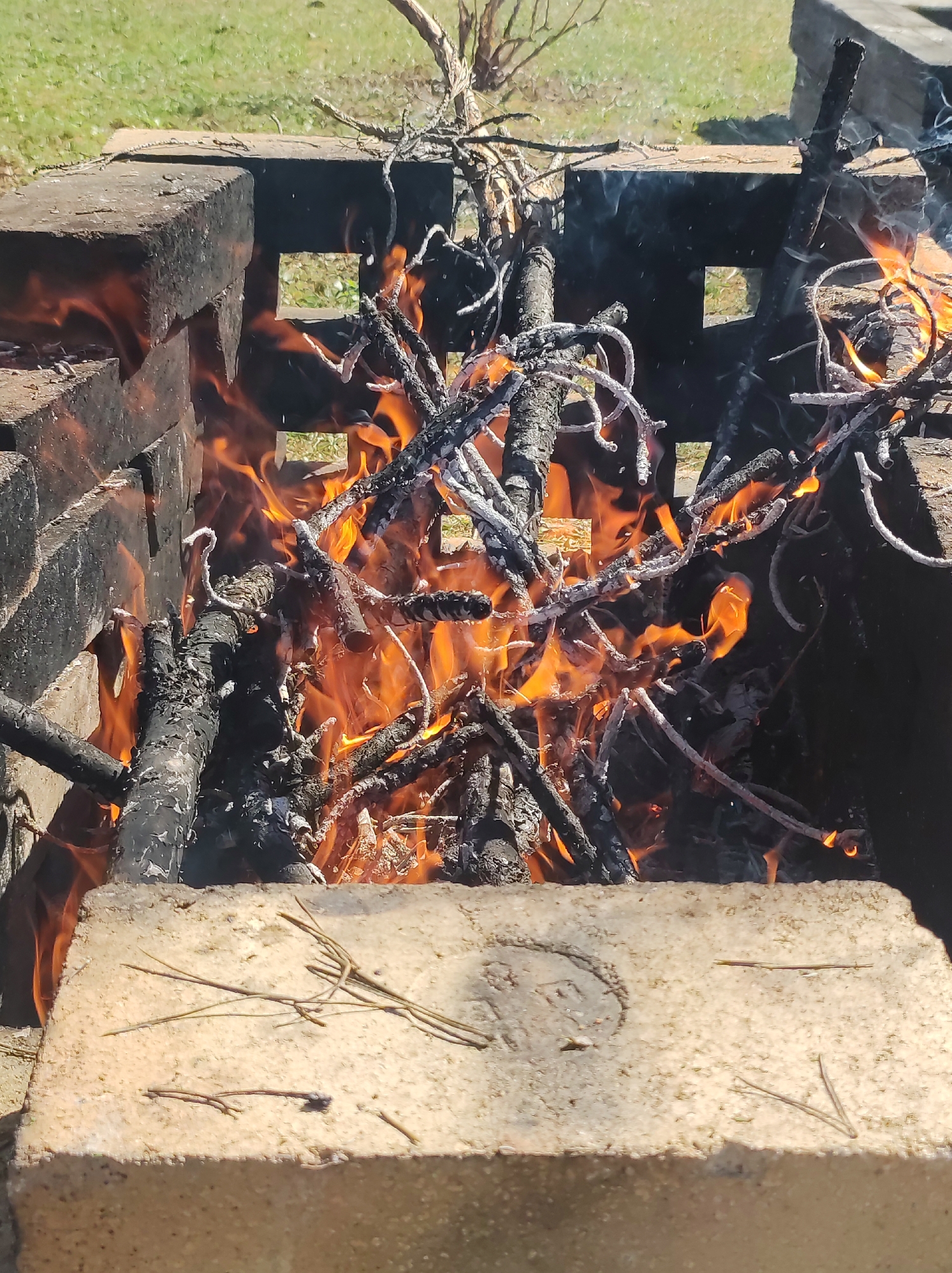 The image depicts a fire burning inside a brick fire pit. The fire is actively consuming branches and sticks, producing flames and smoke. The bricks surrounding the fire are discolored from heat and soot. The surrounding environment appears to be a grassy outdoor area. There is no information about the location in the city of Фоки, Russia.