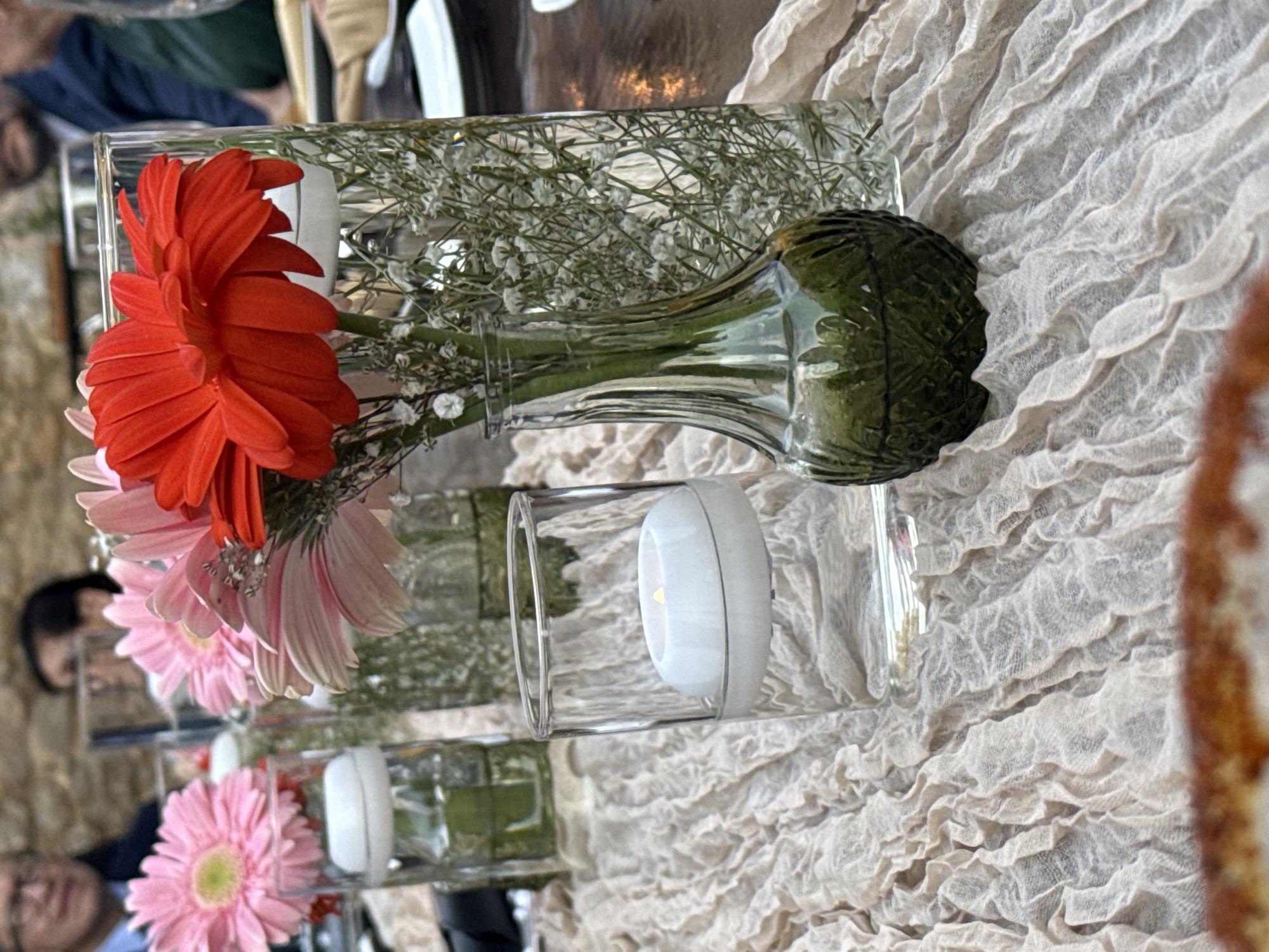 A horizontal close-up shot shows a table setting, likely at a wedding or event, set on a textured beige tablecloth. Three glass vases are positioned on the table, two cylindrical and one shaped like a glass bottle. The cylindrical vases contain white, spherical candles. The glass bottle contains baby's breath flowers and is accented with a large, dark green leaf. Red and light pink gerbera daisies are added near the flowers. In the background, glimpses of other people and place settings can be seen. Based on the stone architecture and decor style, the location could be in Santiago de Querétaro, Mexico.