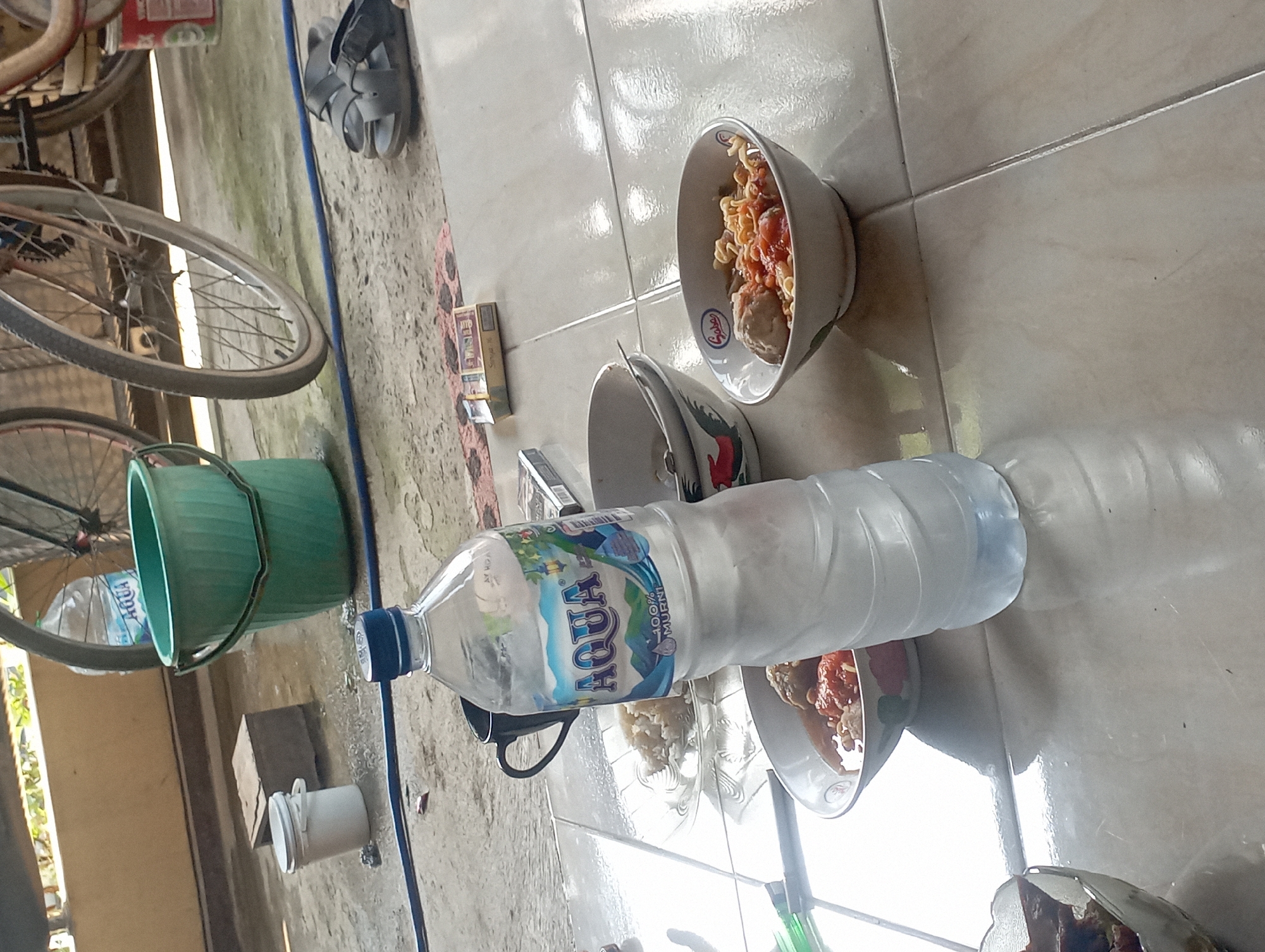 This is a photograph of an outdoor scene in Maos Kidul, Indonesia, on a paved tiled surface. In the foreground, a clear plastic bottle of Aqua brand mineral water stands upright. To the left of the water bottle are two ceramic bowls containing food, one with rice and the other appearing to be a noodle dish with meatballs and sauce. A dark mug is visible behind the water bottle. Further back, a green plastic bucket is present, and to its right, a portion of a bicycle wheel is visible. In the background, a pair of dark sandals are placed on the ground. The scene suggests a casual meal or break being taken outdoors.