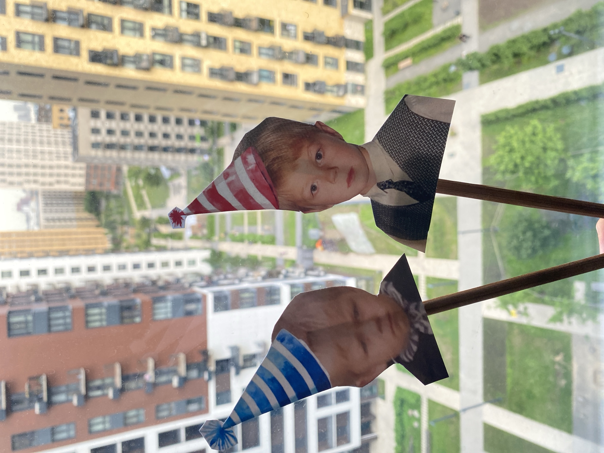 Two collages of children's faces, each wearing a party hat, are mounted on thin wooden sticks, held up against a window. The top collage features a boy with auburn hair, fair skin, and wide, light-colored eyes, wearing a red and white striped party hat and a formal vest with a dark tie. The bottom collage shows another child, with lighter hair and fair skin, also in a blue and white striped party hat. Both collages are placed in front of a window, which offers a view of urban buildings and green spaces.
The background reveals a cityscape with several multi-story buildings, some appearing residential with numerous windows. The buildings have various architectural styles, including a prominent yellow building with horizontal windows in the upper left, and a brick-colored building with darker accents in the lower left. Green park areas with pathways are visible between the buildings, suggesting an outdoor setting with recreational spaces. The lighting suggests it is daytime, with diffused light indicating either overcast weather or the reflection of the sky. The overall impression is a playful, homemade display of children's faces positioned to be seen against an urban backdrop. No text is visible in the image.