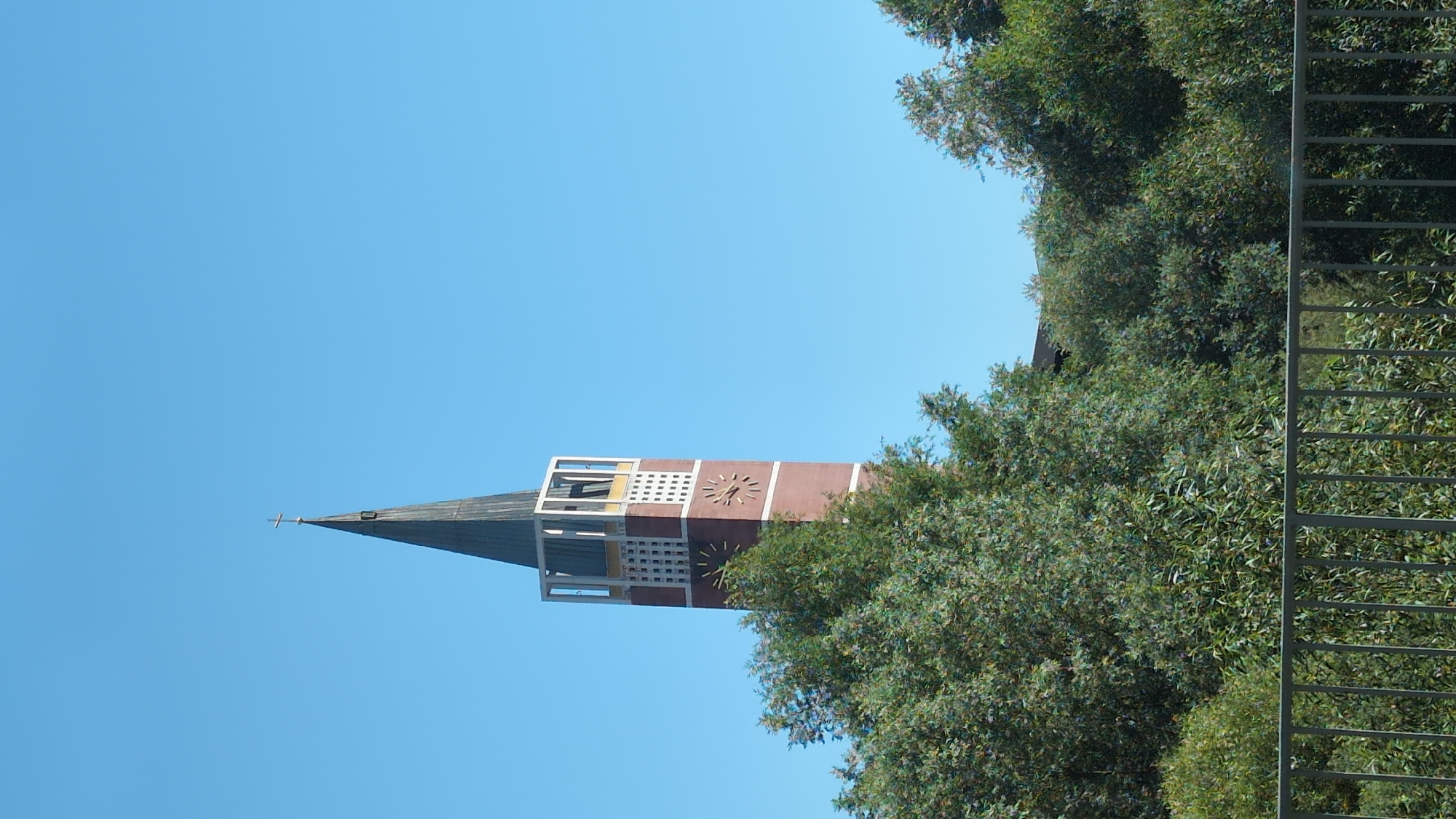 This image, taken on a clear day, shows the tower of the Emmauskirche (Emmaus Church) in Pforzheim, Germany. The tower is tall and slender, with a dark spire topped by a cross. The main body of the tower is constructed of red brick and features a prominent clock face with Roman numerals. Below the clock, there are decorative grid-like elements. Surrounding the base of the tower is lush green foliage, obscuring much of its lower portion. In the foreground, a grey metal fence runs horizontally across the frame. The sky is a uniform, bright blue, with no clouds visible.