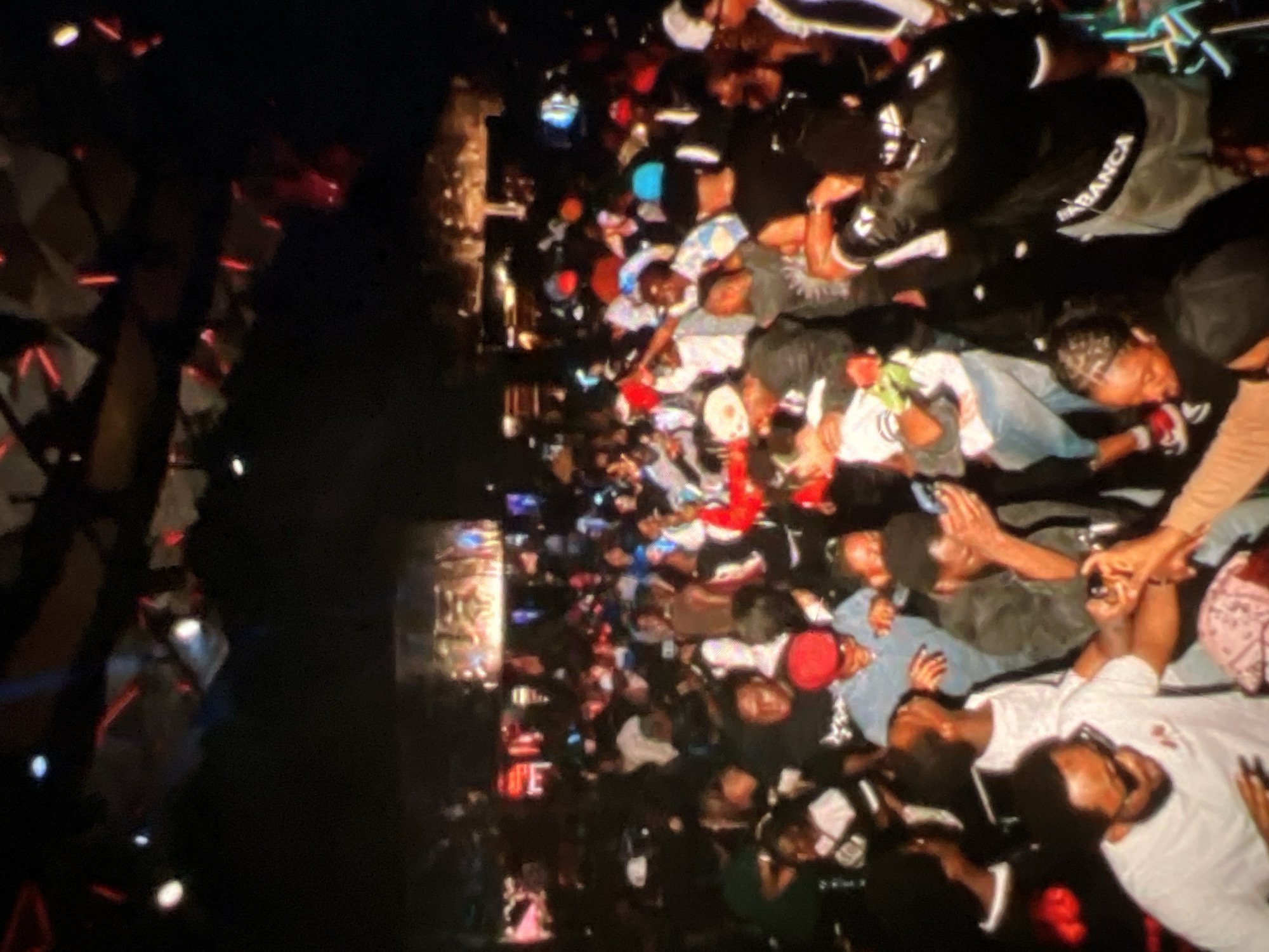 The image depicts a crowded indoor venue, likely a club or concert hall, filled with a large group of people. The lighting is dim with colorful spotlights and stage lighting illuminating the scene, suggesting a lively event is taking place. Many individuals are standing, socializing, and appear to be enjoying themselves. Some are wearing hats and casual attire. In the foreground, several people are visible in close proximity, with one person in a white t-shirt and sunglasses looking towards the camera. Another individual in the foreground is wearing a light blue shirt and a red hat, and seems to be interacting with others. The overall atmosphere appears to be energetic and social. The text "CASABLANCA" is visible on the back of a person's jacket in the mid-ground. The context suggests this event is located in Lagos, Nigeria.