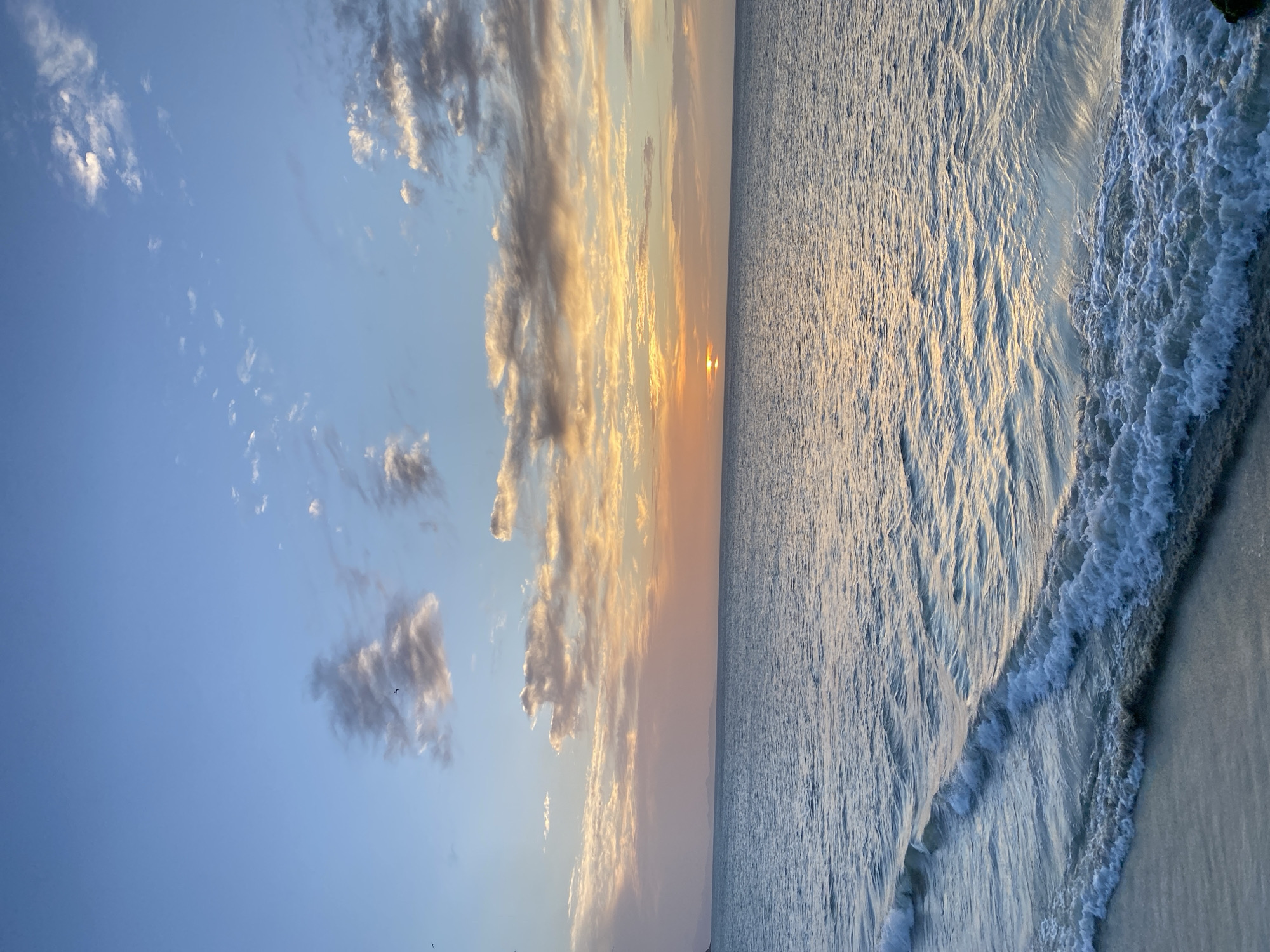 This landscape image shows a sunset scene over the sea in Juan Griego, Venezuela. The sky occupies the left half of the image, a gradient of light blue transitioning to orange and yellow near the horizon. A cluster of clouds, illuminated by the setting sun, is visible towards the center of the sky. The sun itself is setting on the horizon. The sea occupies the right half of the image, its surface textured with light reflecting off the water. A foamy wave is crashing onto the shore in the foreground. The image is taken from a horizontal perspective.