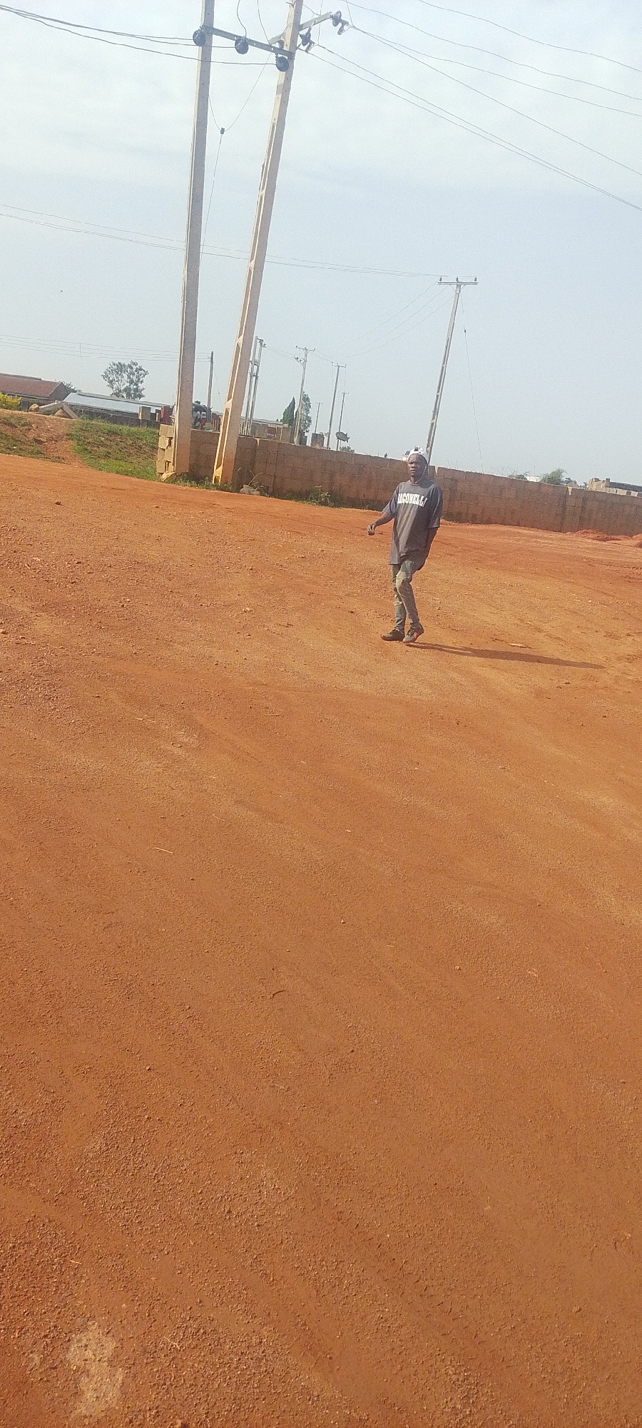 In Jos, Nigeria, a man walks on a dirt road. He is wearing a grey t-shirt with white lettering and blue jeans. He has a white cap on his head. He is walking away from the camera, towards the right side of the frame. The road is made of red dirt and is uneven. In the background, there are power poles and a brick wall. The sky is overcast.