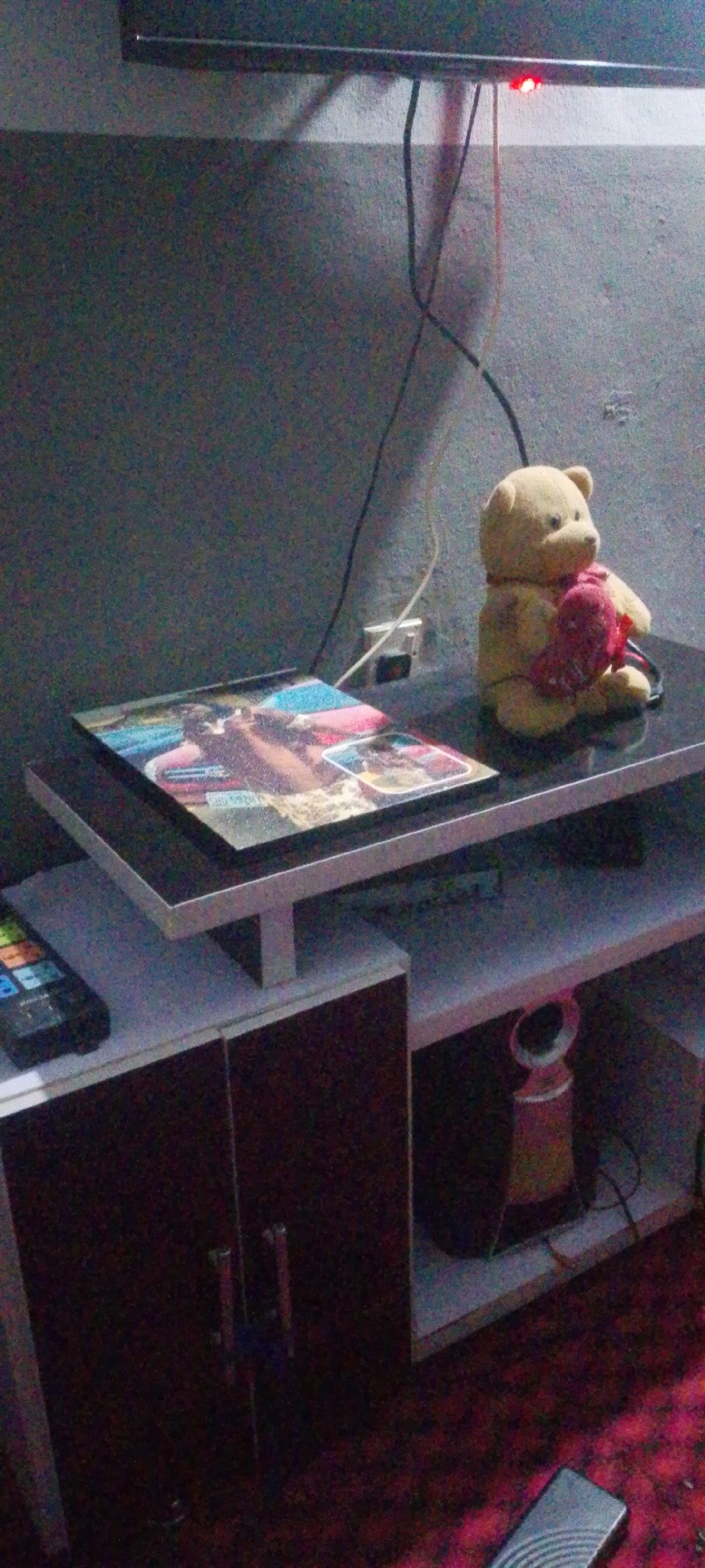 The image shows a close-up of a TV stand in an indoor setting. A framed picture or a magazine with an image of a person and colorful abstract shapes is placed on the top shelf. Next to it, a plush teddy bear is seated, holding a pink heart. A black remote control is visible on the left side of the shelf. The TV stand itself appears to be made of wood and has a dark brown cabinet with two doors at the bottom. A pink and orange cylindrical object, possibly a speaker or a decorative item, is on the lower shelf. Wires are visible in the background, connected to an electrical outlet. The wall behind the TV stand is a dark gray. The floor is covered with a red carpet. The lighting is dim, suggesting it might be evening or nighttime. The location is specified as Unknown, Unknown.