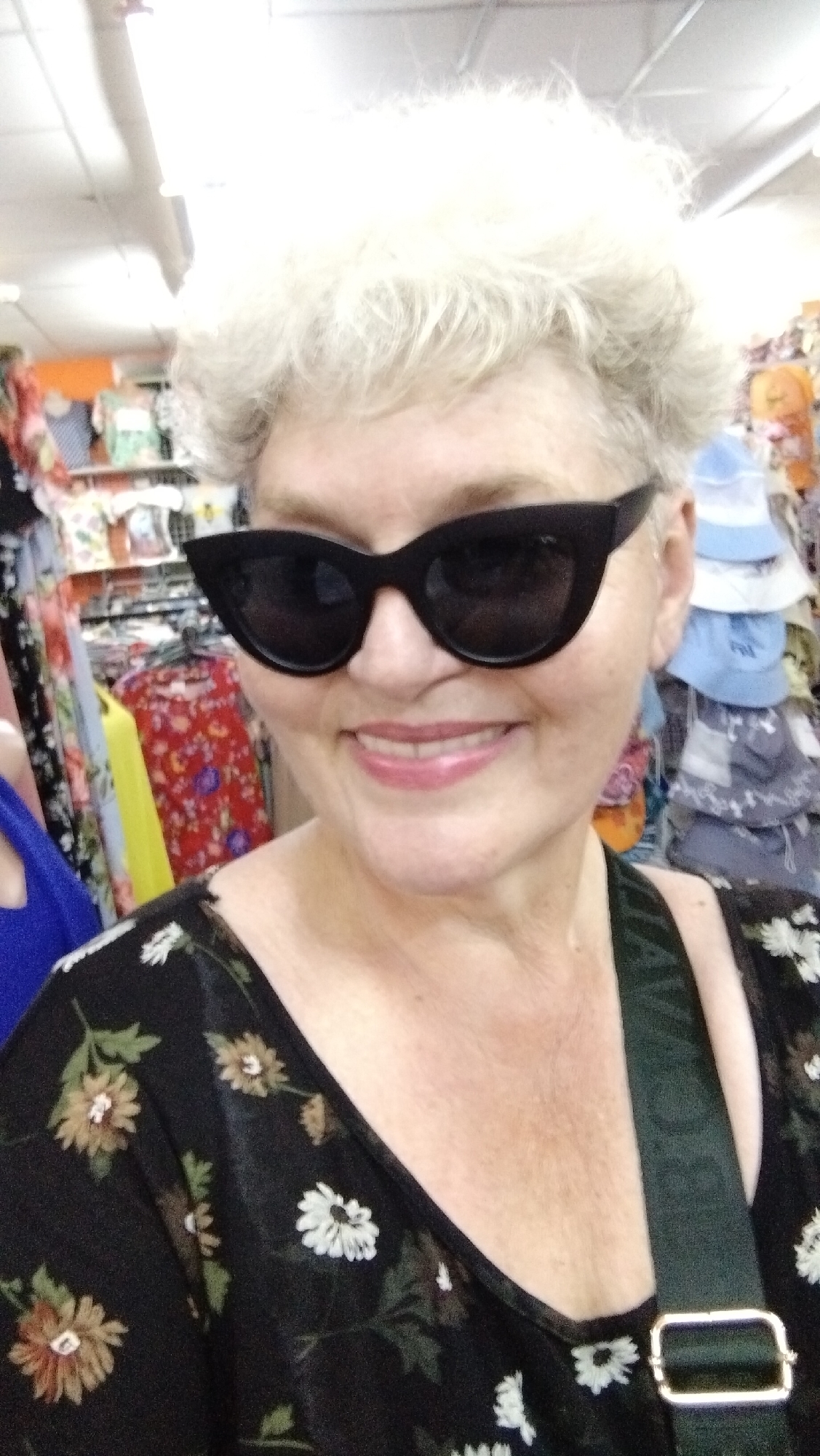 The image shows a woman with short, white, curly hair, wearing large black sunglasses and a black dress with a floral pattern. She is smiling and appears to be indoors, possibly in a store, with racks of clothing visible in the background. The context of Horishni Plavni, Ukraine is not evident from the visual information alone.