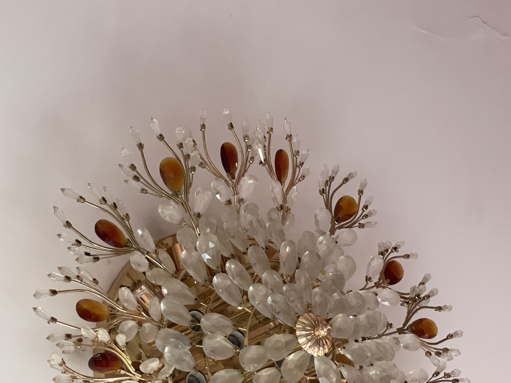 The image shows a close-up of a decorative light fixture, likely a chandelier, attached to a ceiling. The fixture is elaborate, featuring numerous clear, teardrop-shaped crystals and smaller, faceted clear crystals arranged in a floral or fan-like pattern. Interspersed among the clear crystals are teardrop-shaped amber or dark brown stones. The metal components of the fixture, including the branches holding the crystals and a central decorative element at the base, appear to be gold-toned. The ceiling is a pale, uniform color, possibly white or a very light shade of pink, with a slightly textured surface. There is an area of discoloration or damage visible on the ceiling to the right of the fixture, resembling a water stain or peeling paint. The photograph is taken from a slightly upward angle, focusing on the fixture itself. The image does not contain any people, animals, or explicit indications of the city of Sochi, Russia.