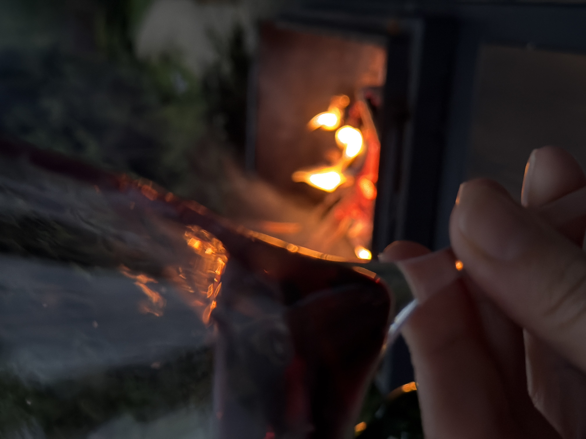 A close-up image shows a hand holding a clear glass, likely a wine glass, filled with a dark liquid in front of a wood-burning stove or fireplace. The fire inside the stove is visible as bright orange and yellow flames, and its reflection is distorted in the glass. The background appears dark, with indistinct shapes that may be foliage or other objects. The location, Zimyonki, Russia, is not definitively verifiable from the image content.