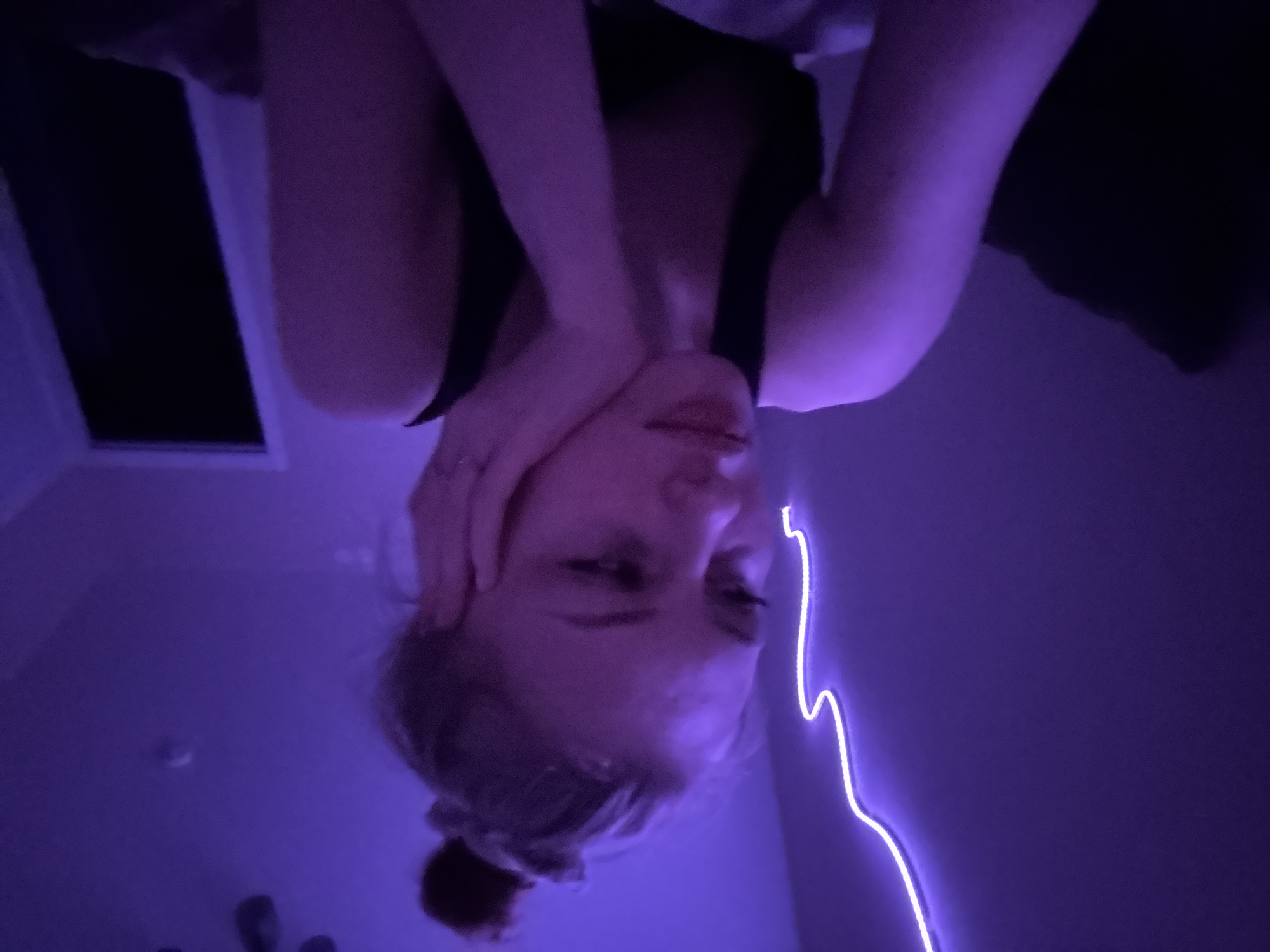 A low-angle shot shows a woman's face and shoulders illuminated by a purple light. Her hair is pulled back in a bun. She is wearing a dark tank top. Her left hand is positioned near her cheek. A window is visible in the upper-left corner of the shot, and a glowing purple light runs along the wall to the right. The scene appears to be indoors. The exact location in Perm, Russia cannot be determined from the provided image.