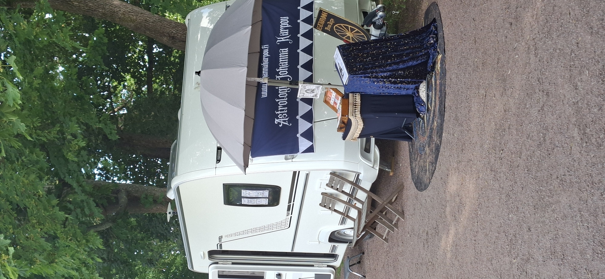 This image depicts an outdoor setup featuring a white caravan, likely serving as a mobile business. In front of the caravan, a table is draped with a dark blue, glittery fabric and covered with a dark blue cloth, on which rests an open suitcase and a book. A large grey umbrella is open and positioned above the table, providing shade. A dark blue banner with white lettering is hung on the caravan, reading "Astrologi Johanna Karpou" and a website address. To the left of the caravan, two wooden folding chairs are placed on the ground. The ground itself is covered in gravel. Behind the caravan, trees with lush green foliage are visible, suggesting a natural or park-like setting. The overall scene appears to be a temporary stall or display for an astrology service, potentially at an event or market. Without further information, the specific location in Turku, Finland, cannot be determined from the image alone.