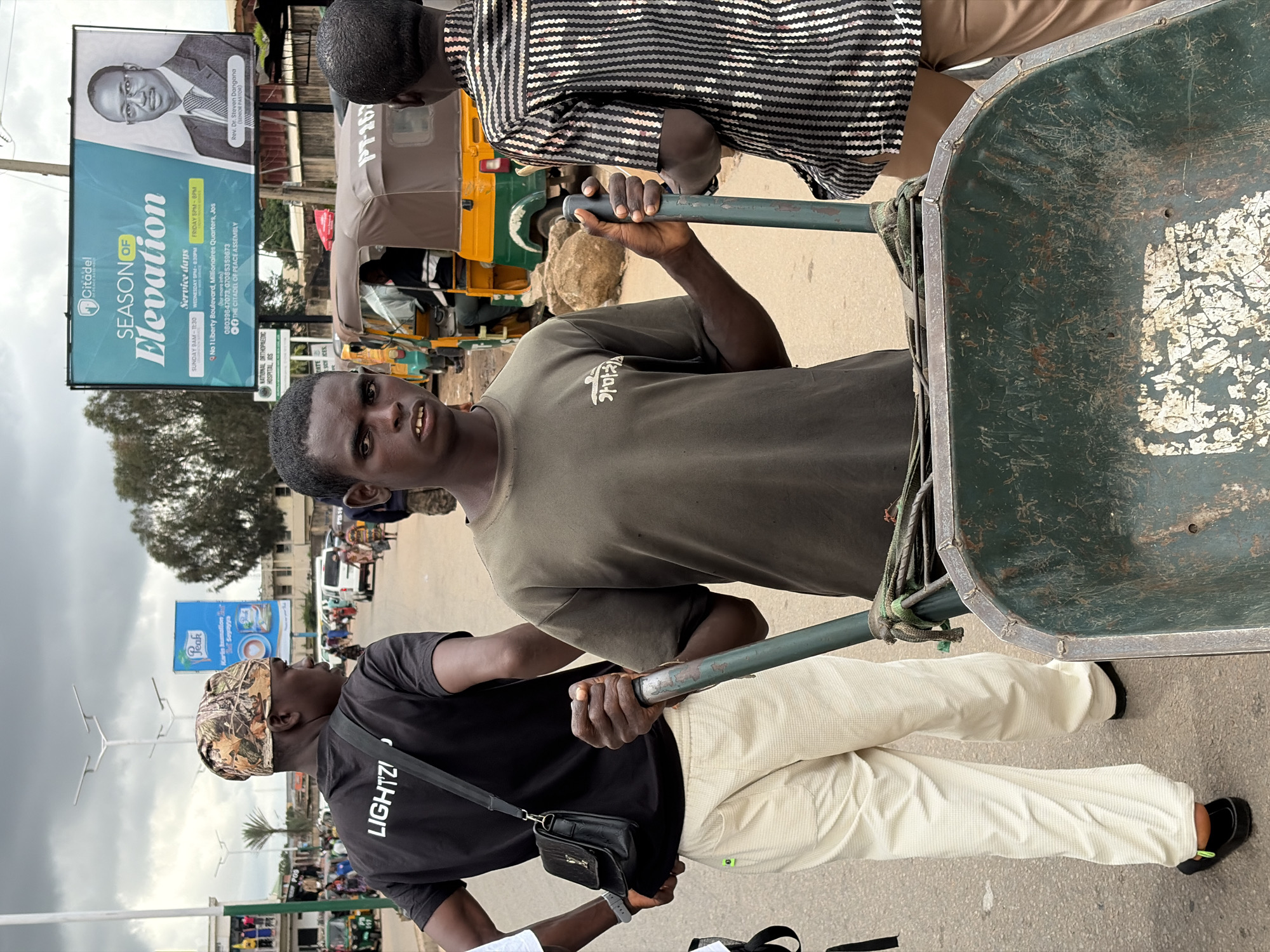 The image captures a street scene in Jos, Nigeria, with a young man in the foreground pushing a weathered green wheelbarrow. He wears a dark green t-shirt with a faint white symbol on the left chest. His expression is serious as he looks towards the camera. Behind him, another young man in a black t-shirt with "LIGHTZI" printed in white lettering is walking away, carrying a black bag over his shoulder. He wears a camouflage durag and light-colored pants. To the right of the wheelbarrow pusher, a third person is visible from the back, wearing a black and white striped shirt and tan pants. In the background, several auto-rickshaws are parked and moving, along with other pedestrians and vehicles. A large billboard advertising "Citadel: Season of Elevation" featuring a portrait of a man in a suit is prominently displayed overhead. The environment appears to be a busy urban street with buildings and streetlights visible. The overall atmosphere suggests an active marketplace or thoroughfare.