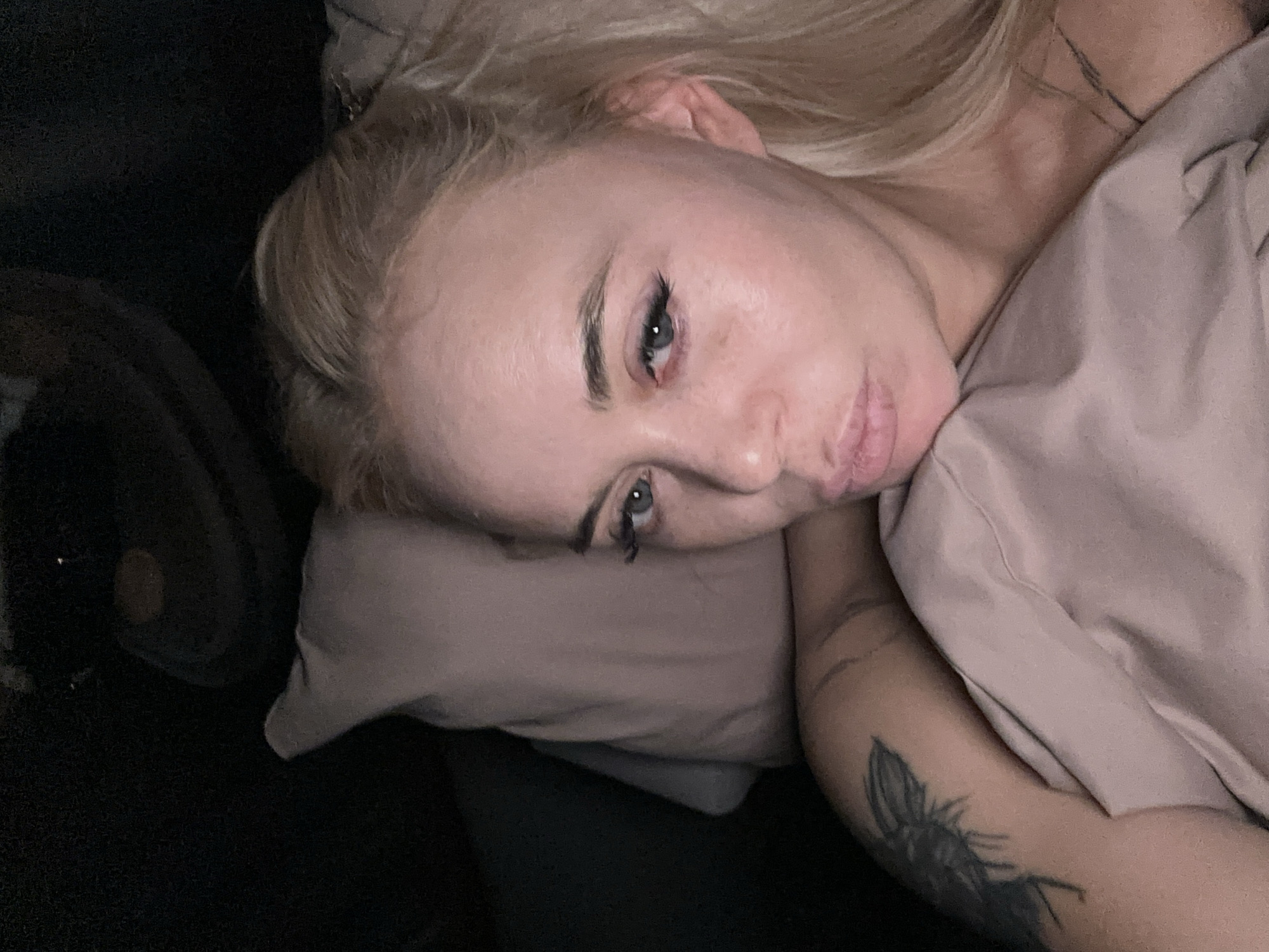 A woman with blonde hair is lying on a bed with a pillow and a blanket. She has a tattoo on her arm. The location is Высокий, Russia.
