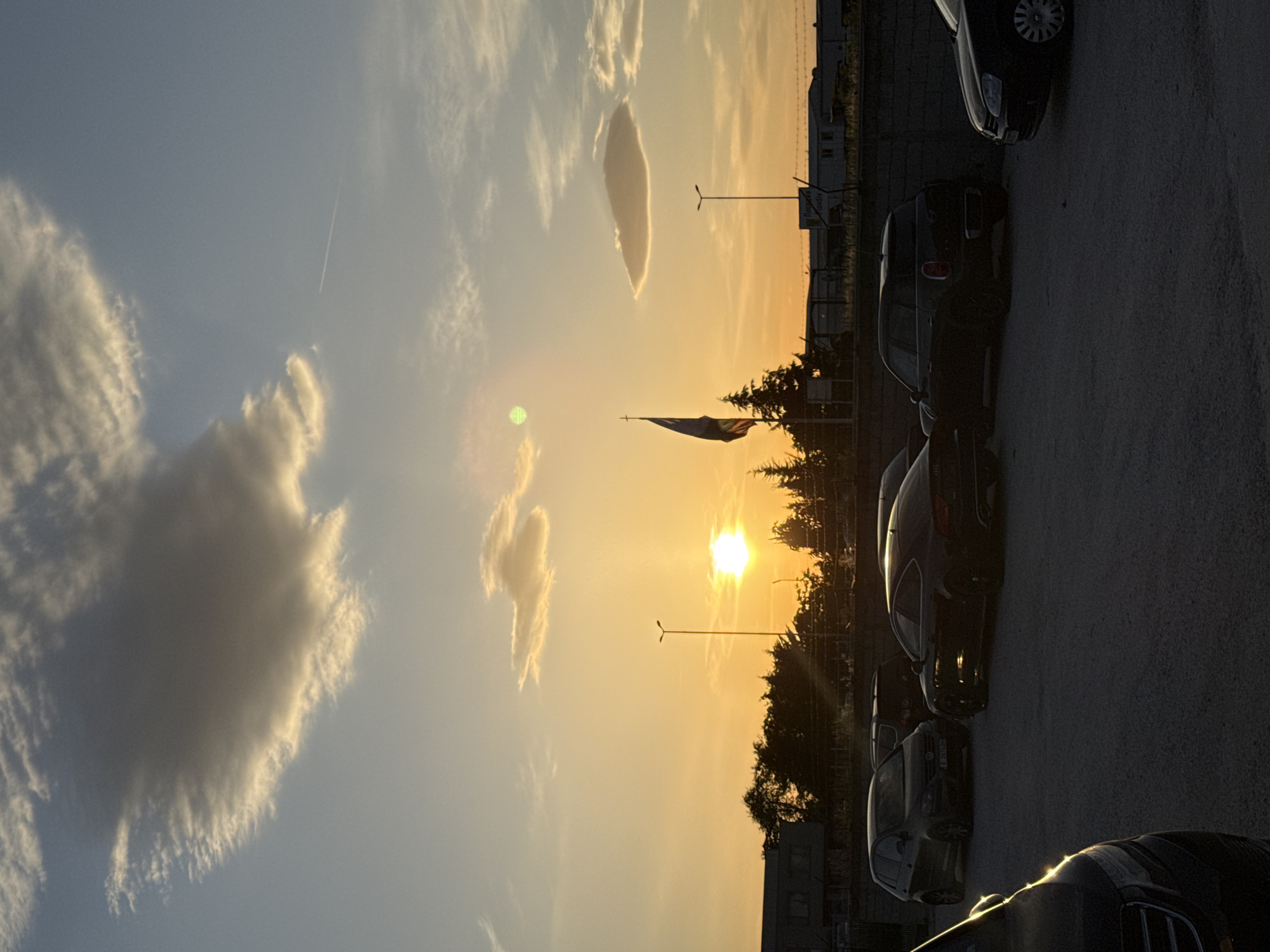 The image captures a sunset scene in an urban environment, likely a parking lot on the outskirts of an unidentified city. Several cars of varying models and colors are parked along the right edge of the frame. Behind the cars, there is a treeline silhouetted against the setting sun, with buildings and utility poles visible further back. A flag is also seen amidst the trees. The sky is filled with clouds, some illuminated by the sunlight, creating a gradient of colors from blue to orange and yellow. A contrail is visible in the upper left part of the sky.