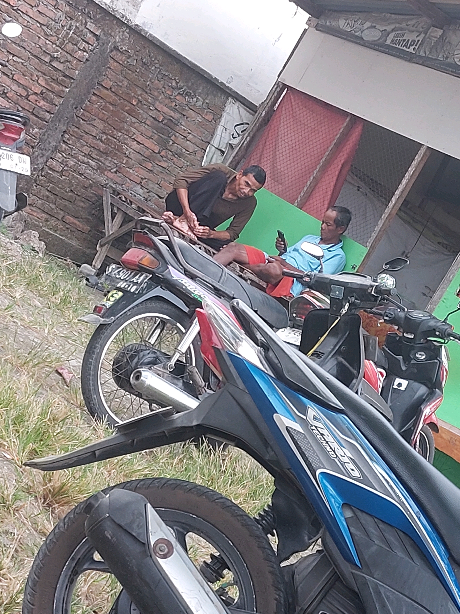 In SARANGAN, Indonesia, two adult males are present in an outdoor scene. The man on the left, wearing a brown shirt and dark pants, is seated with his feet on a wooden structure, appearing to be getting a foot massage. The man on the right, dressed in a light blue shirt and red shorts, is sitting on a motorcycle and looking at his mobile phone. They are positioned next to several parked motorcycles, including a black one with a license plate that appears to read "S 4901 AA," and a red one. The background features a brick wall, a building with red mesh curtains and a banner with Indonesian text, and overgrown grass. The overall environment suggests a casual outdoor gathering or waiting area.